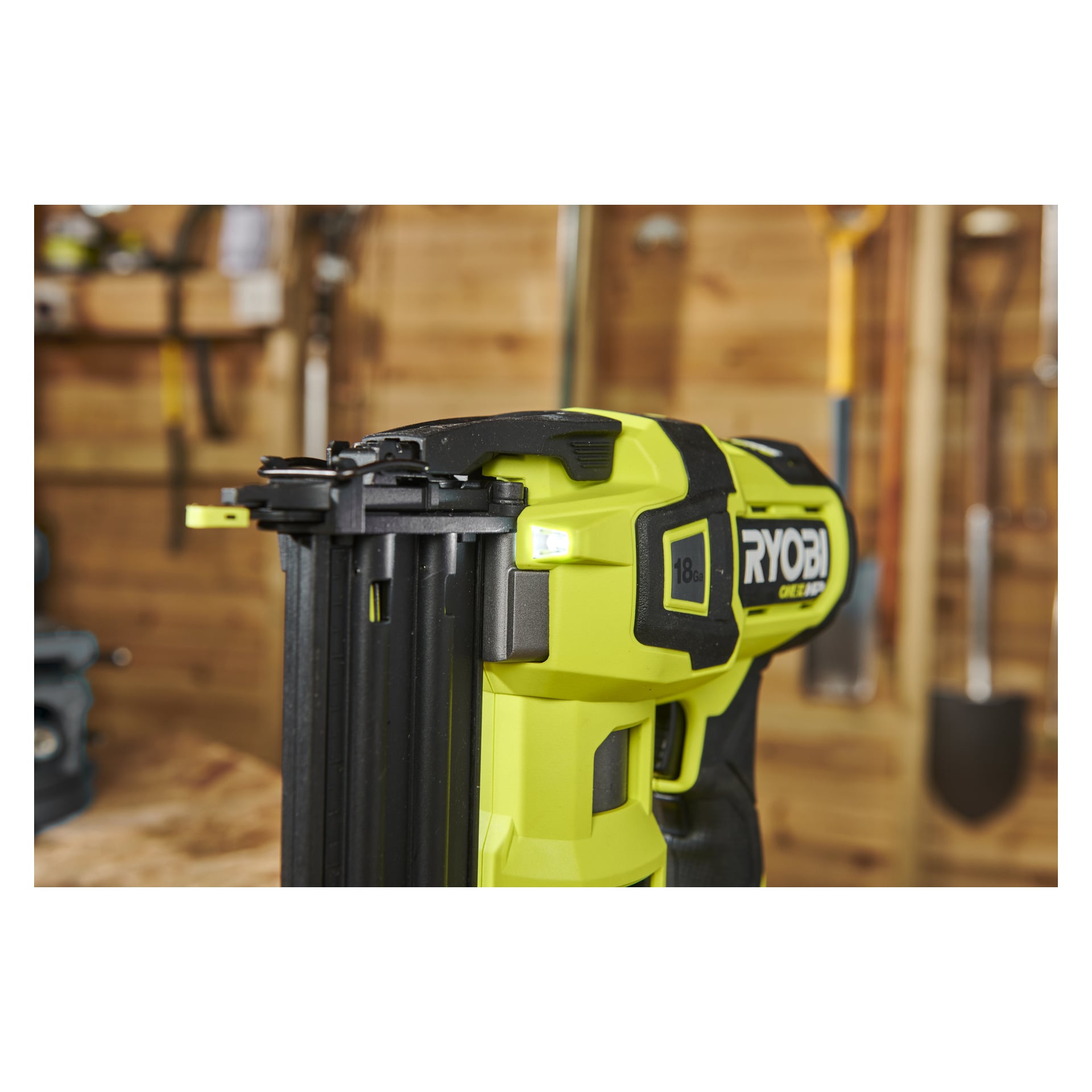 Grön Ryobi One+ HP Brad spikpistol AirStrike R18GN18X-0 visades på nära håll på en arbetsbänk, med olika träverktyg och en spade hängande på väggen i den mjukt fokuserade bakgrunden.