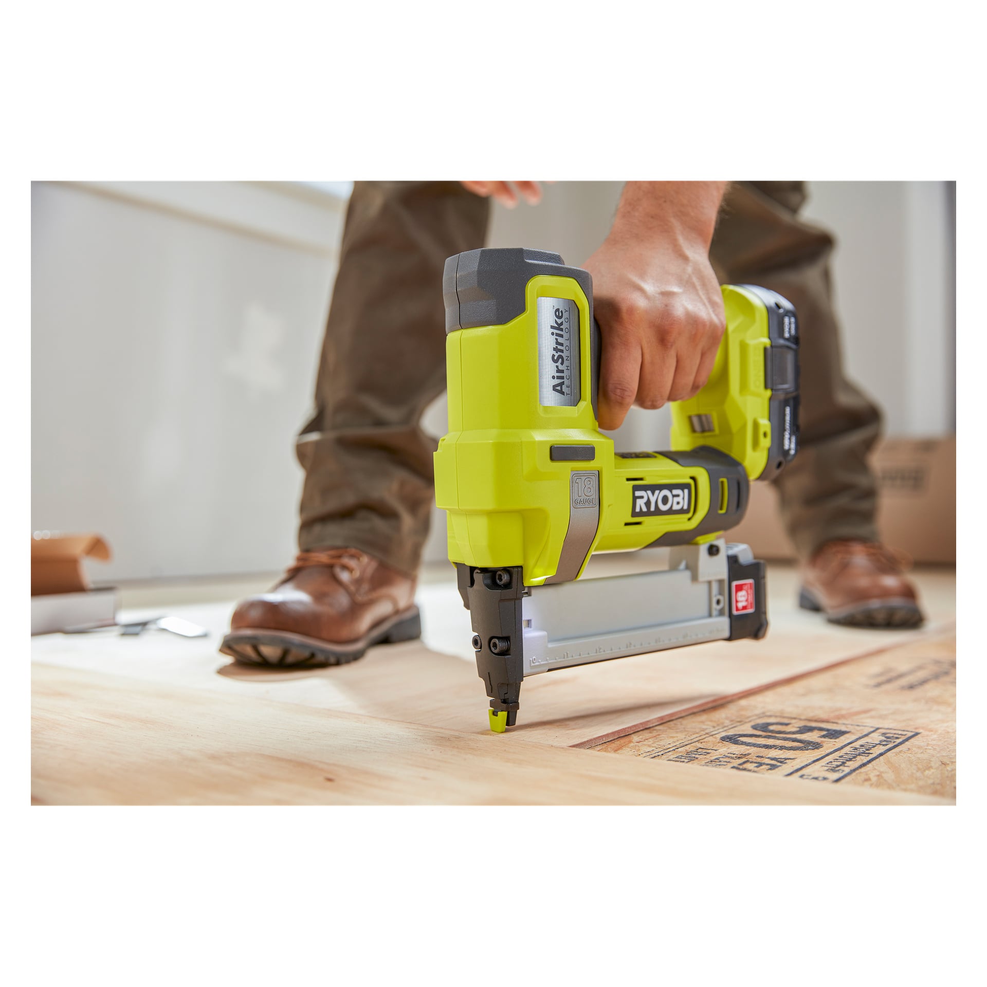 Någon som bär bruna arbetsskor använder en Ryobi One+ HP Brad Nailer AirStrike R18GN18X-0 för att fästa plywoodgolv i en inomhusarbetsplats.