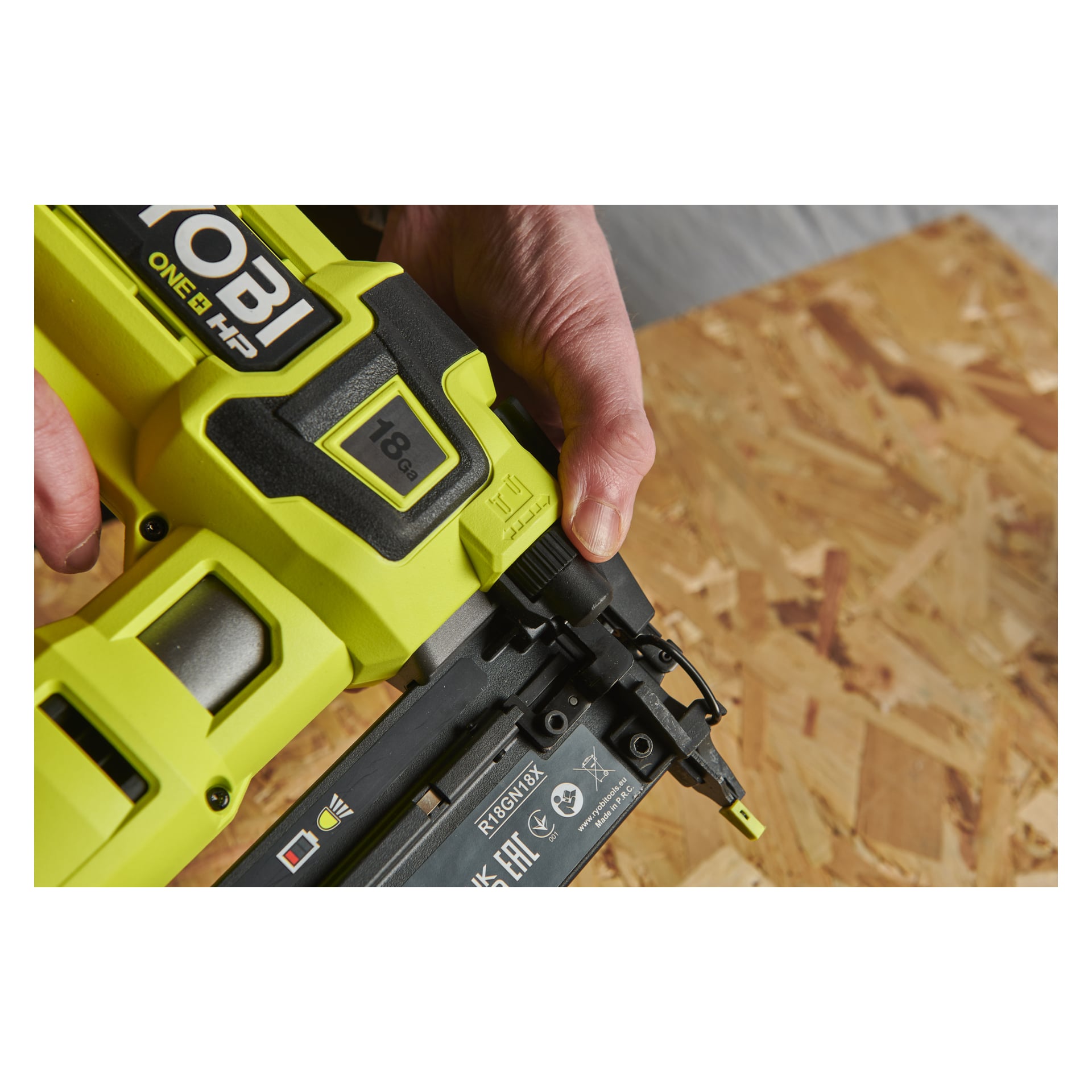 Någon håller i en grön Ryobi One+ HP AirStrike R18GN18X-0 spikstift och placerar den ovanför en sektion av orienterad strandbräda (OSB) redo för användning. Både verktyget och individens händer är tydligt synliga.