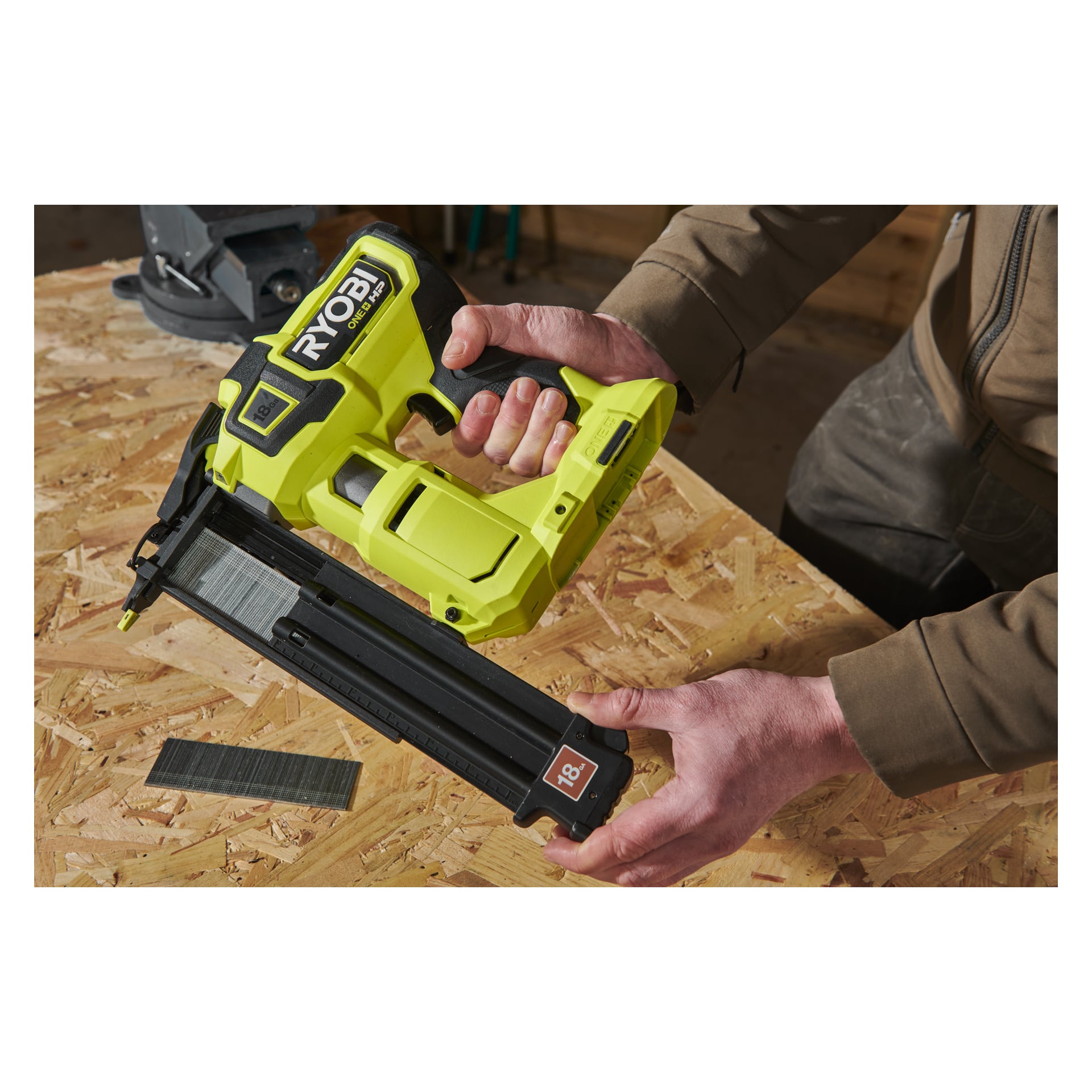 Någon sätter in spikar i en grön Ryobi One+ HP AirStrike R18GN18X-0 spikmaskin på en träarbetsbänk. En smal remsa av spikar vilar bredvid verktyget, och en del av personens bruna ärm kan ses.