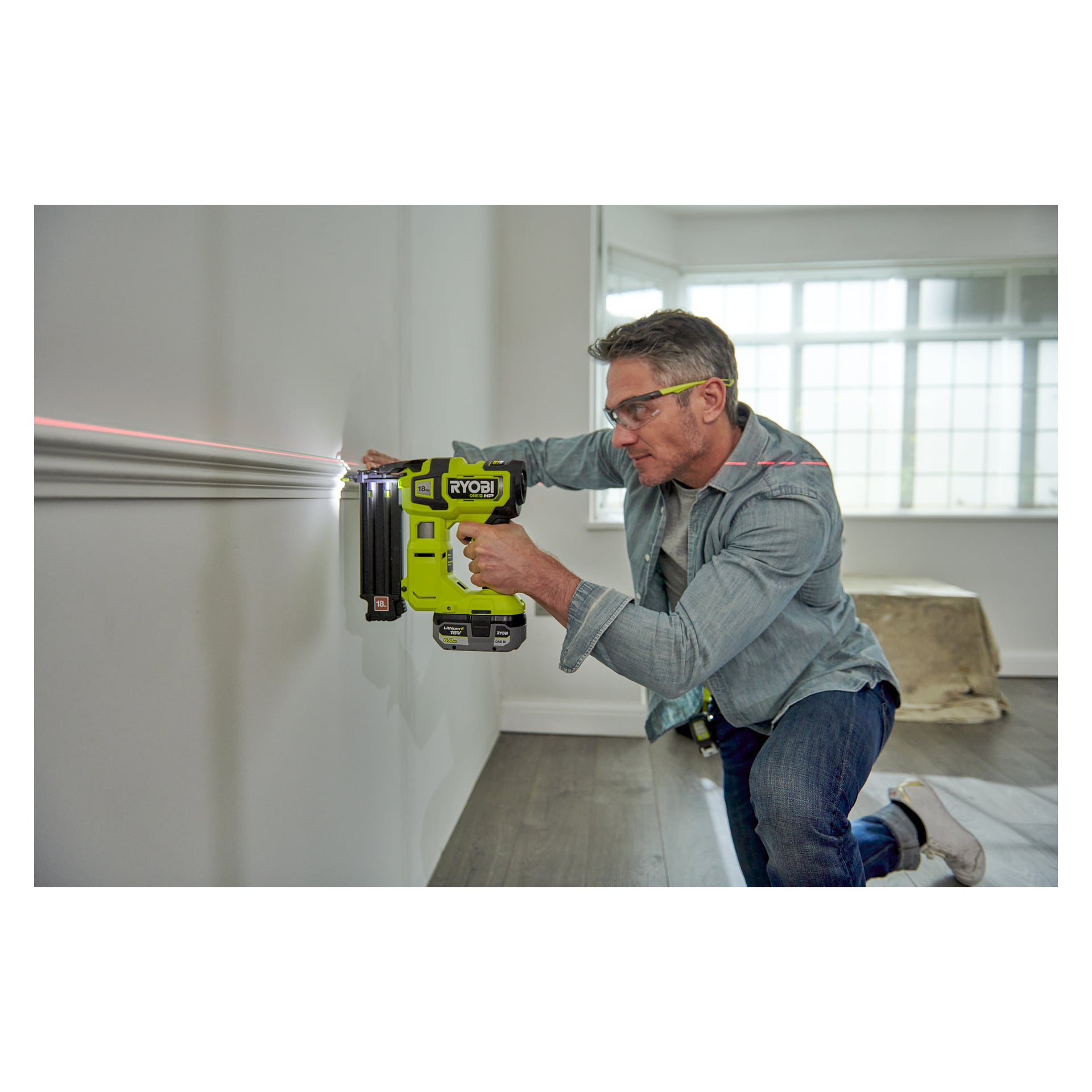 Med skyddsglasögon använder man en grön Ryobi One+ HP AirStrike spikpistol (modell R18GN18X-0) för att fästa lister på en vägg, enligt en röd lasernivålinje i ett starkt upplyst rum med stora fönster.