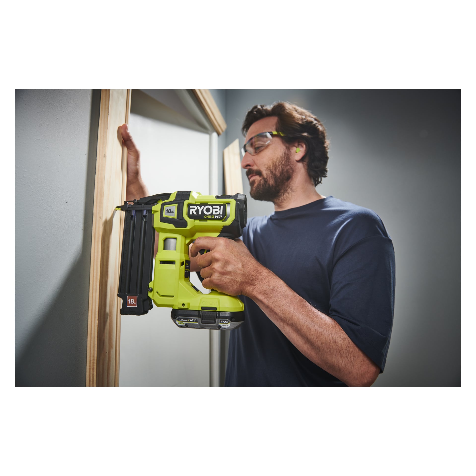 En person som bär skyddsglasögon använder Ryobi One+ HP Brad Nail Gun AirStrike R18GN18X-0 för att fästa en träram på en dörröppning i ett väl upplyst rum med grå väggar.