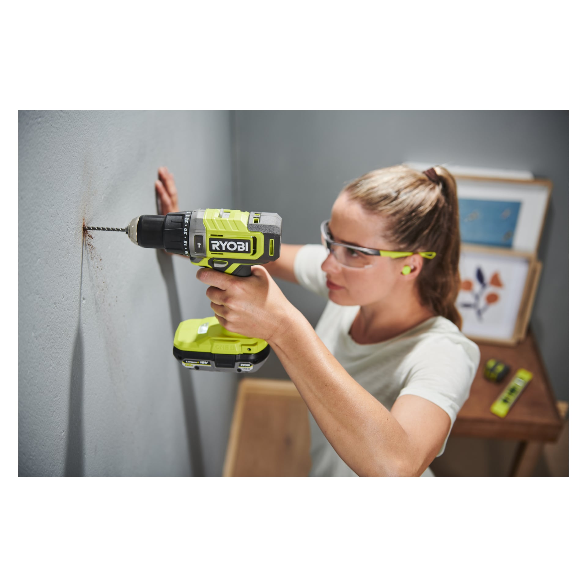 En person utrustad med skyddsglasögon och öronproppar använder en Ryobi One+ Impact borrmaskin RPD18-0 och borrar in i en grå vägg. I bakgrunden finns inramade konstverk och ett grönt måttband.
