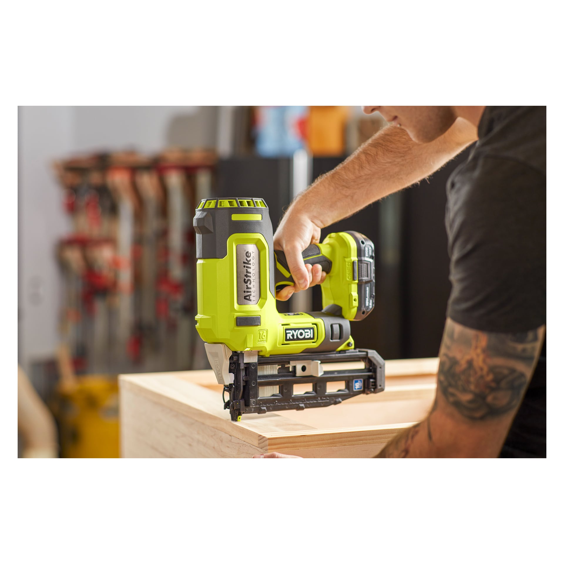 Jemand befestigt Holzstücke in einer Werkstatt mit einem grünen Ryobi One+ R16GN18-0 kabellosen Brad-Nagel, während im Hintergrund verschiedene Werkzeuge und Klemmen unscharf sichtbar sind.