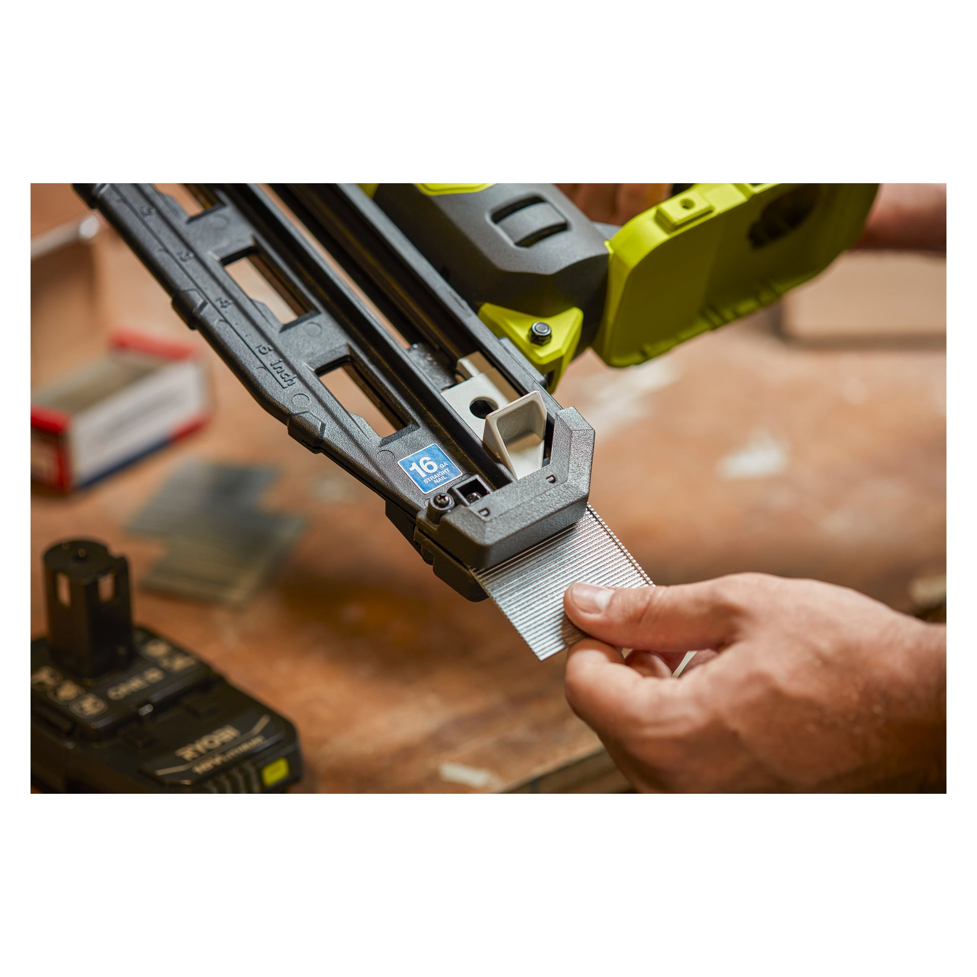 Eine Person setzt einen Streifen 16-Gauge-Nägel in eine Ryobi One+ Brad-Waffe R16GN18-0 auf einer Werkbank, wobei ein Batteriepack und eine Nagelbox in der Nähe platziert sind, was auf die Vorbereitung für Holzbearbeitung oder Bau hinweist.