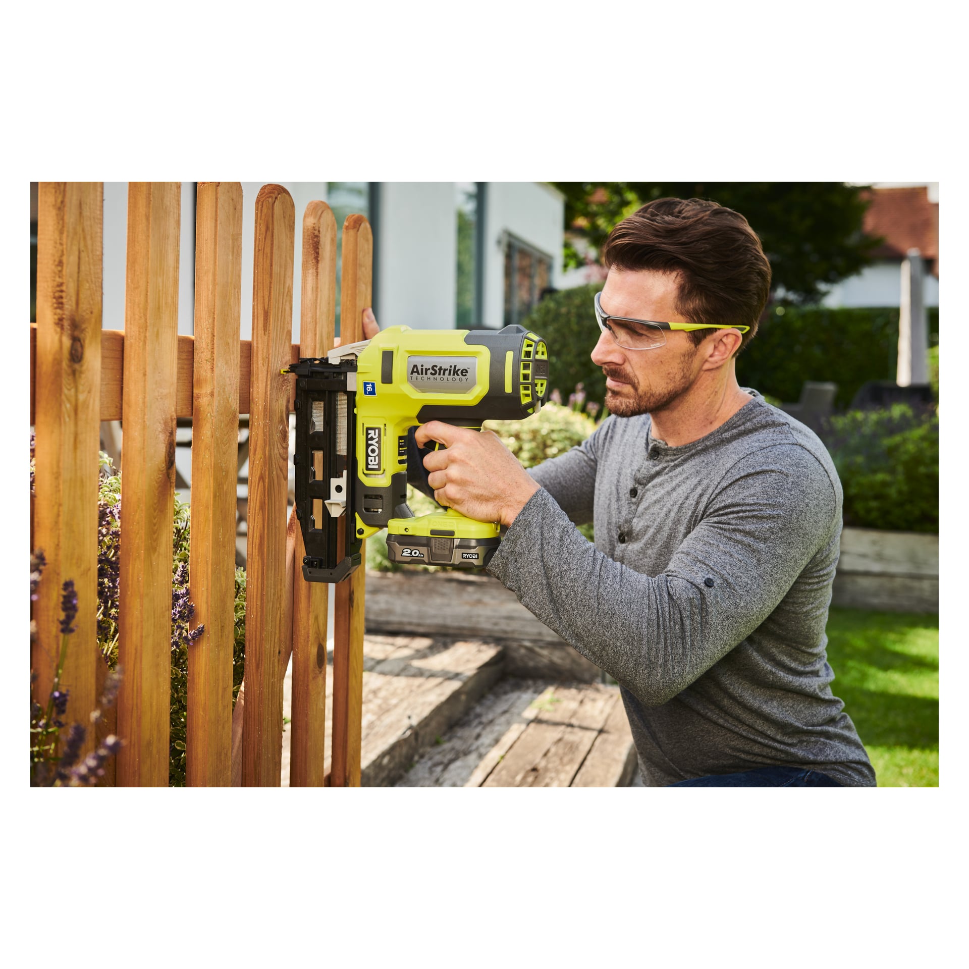 Ein Mann mit Sicherheitsbrille kniet draußen und konzentriert sich und benutzt eine Ryobi One+ Brad-Pistole R16GN18-0, um eine Holzzaunlatte zu befestigen.