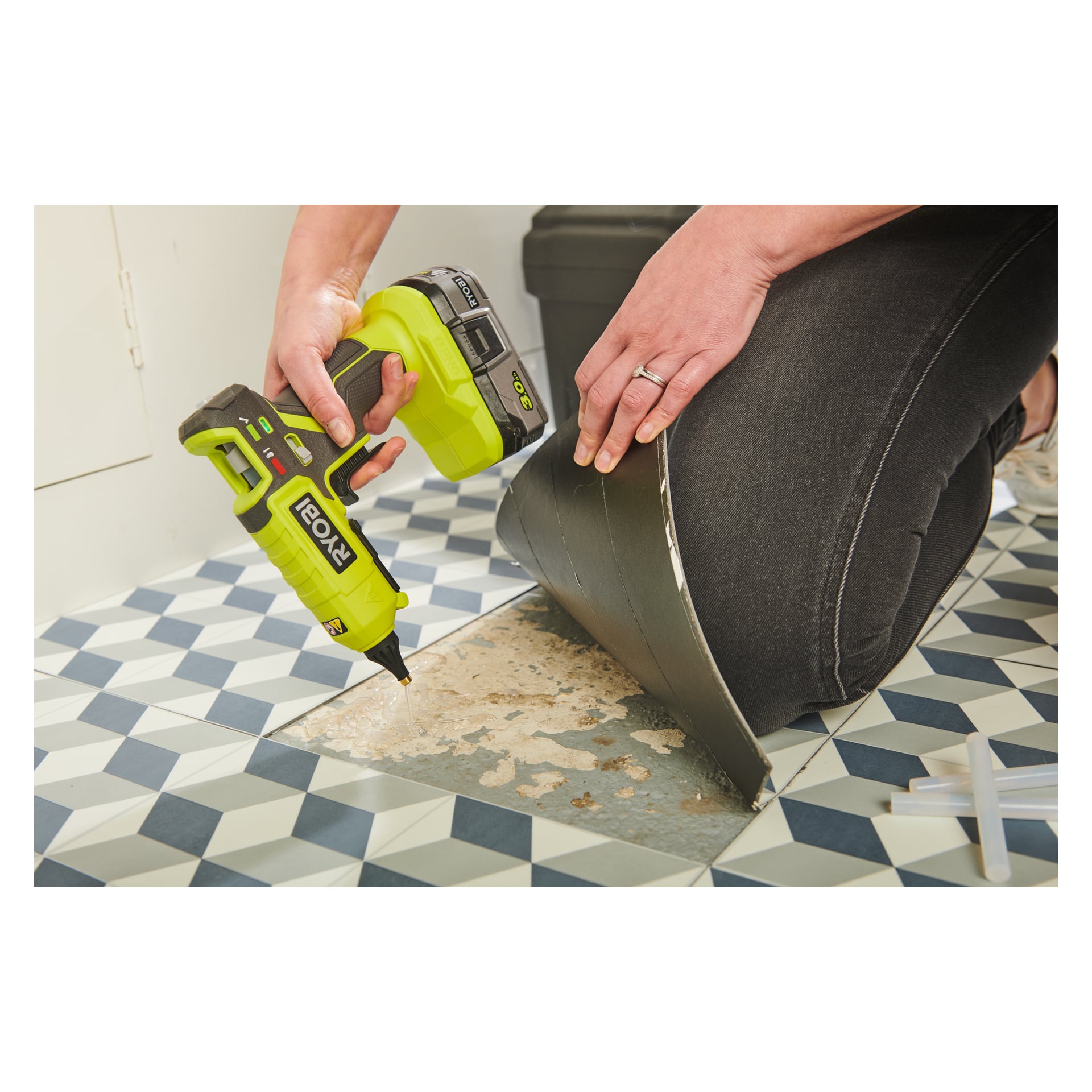 En person knäböjer på geometriska plattor, håller en Ryobi One+ batteridriven limpistol (RGL18-0) i ena handen och drar bort mönstrat vinylgolv med den andra, vilket avslöjar det bara golvet under.