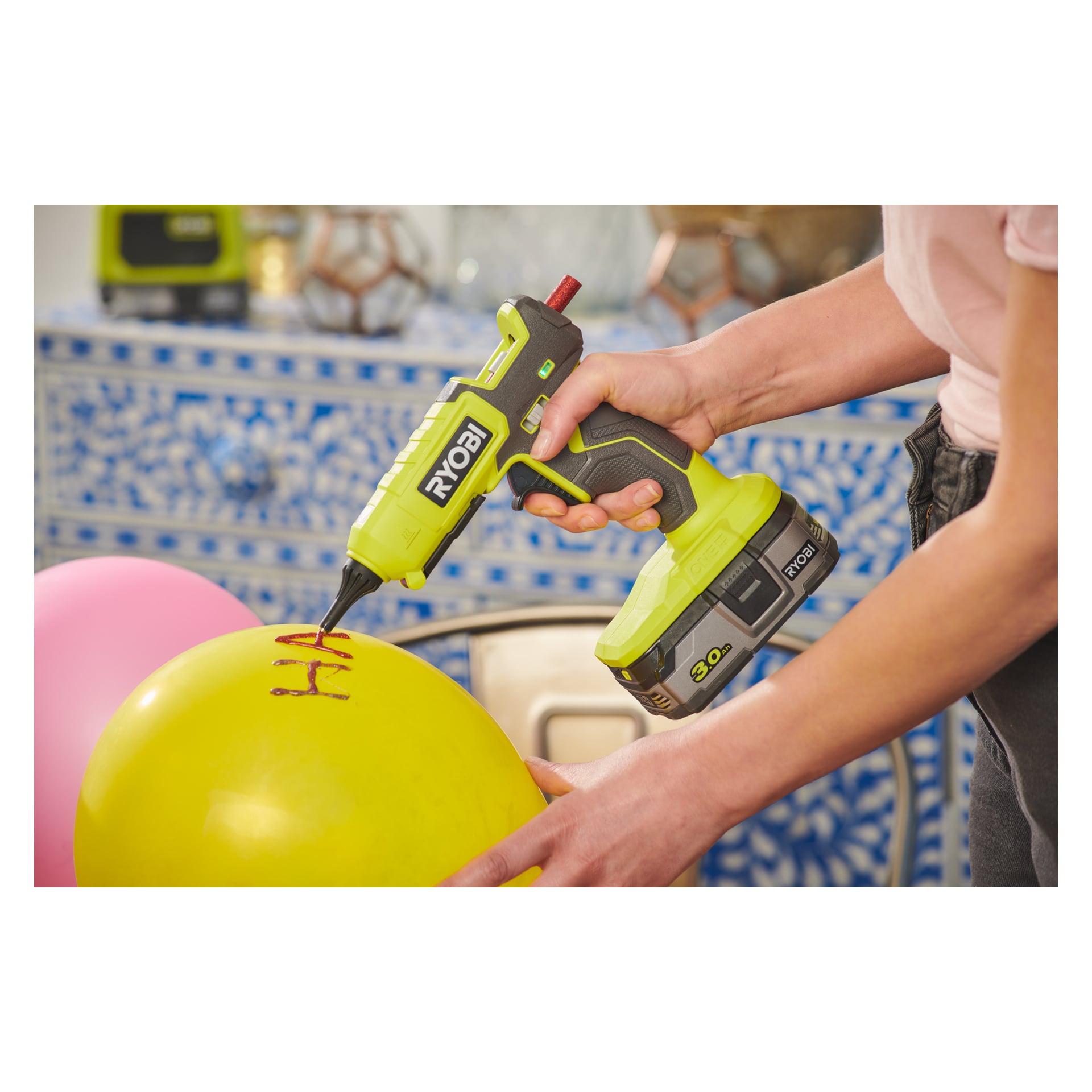 En person använder en Ryobi One+ Battery-driven limpistol RGL18-0 för att skriva med rött lim på en gul ballong, medan en rosa ballong och mönstrade blå och vita plattor kan ses i bakgrunden.
