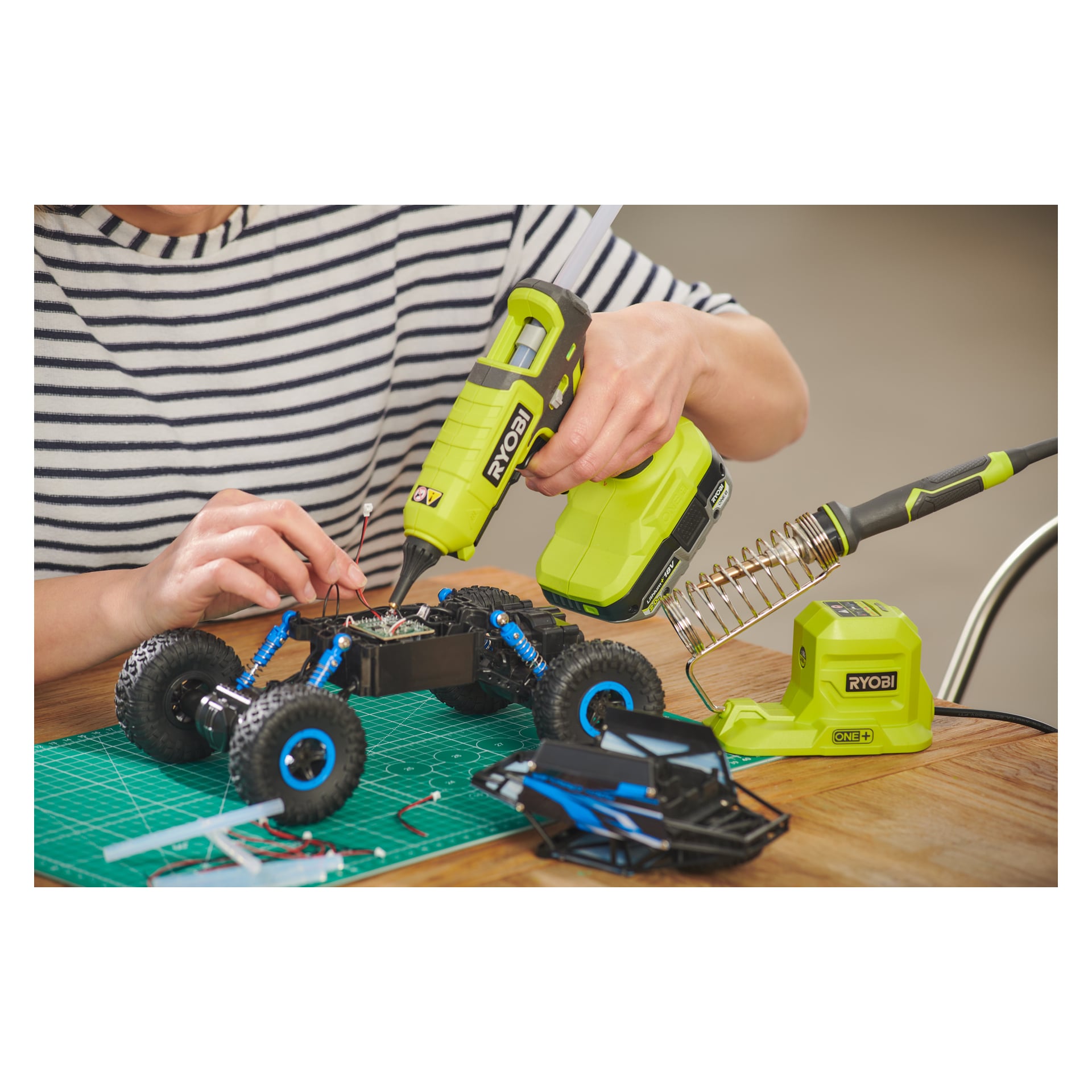 Med en randig skjorta använder en person en Ryobi One+ batteridriven limpistol RGL18-0 för att reparera ledningarna i en fjärrstyrd bil. Olika verktyg och bildelar ligger utspridda på en grön skärmatta.