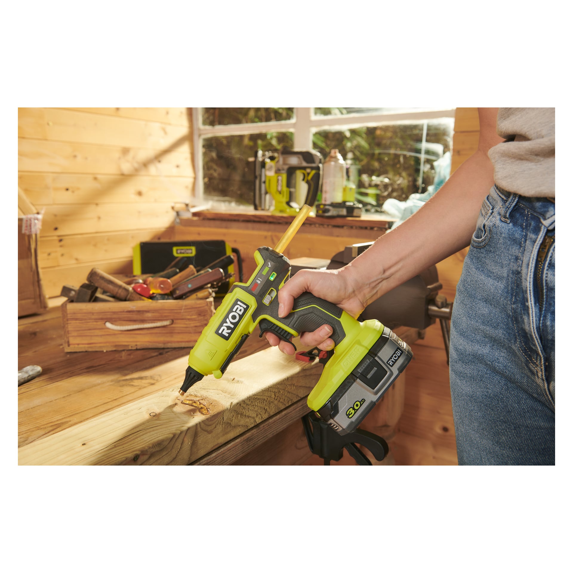 En person i jeans använder en Ryobi One+ batteridriven limpistol RGL18-0 på en träarbetsbänk inne i en verkstad. Olika verktyg och en verktygslåda står på bänken, och solljuset strömmar in genom ett fönster bakom dem.