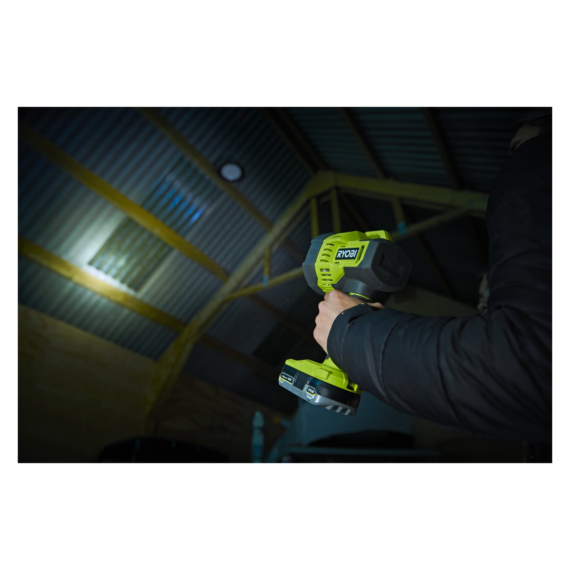 Någon som bär en svart jacka håller upp en grön Ryobi One+ Portable LED-strålkastare RLS18-0 och riktar dess ljus mot metalltaket i ett svagt upplyst skjul eller lada.