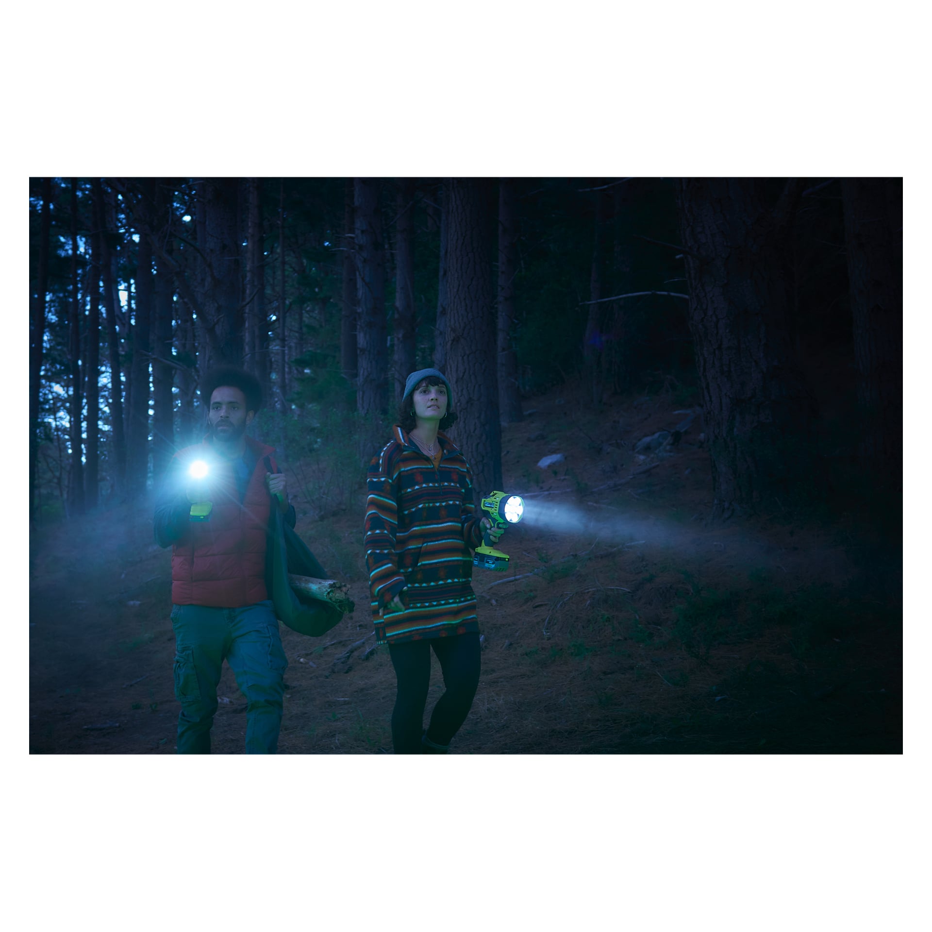 Två personer går genom en mörk skog, båda bärande en Ryobi One+ Portabel LED-pannlampa RLS18-0. Personen till vänster bär en röd väst och cargobyxor, medan den till höger har en randig tröja och mössa. Båda använder sina pannlampor för att lysa upp stigen framför sig när de rör sig framåt.