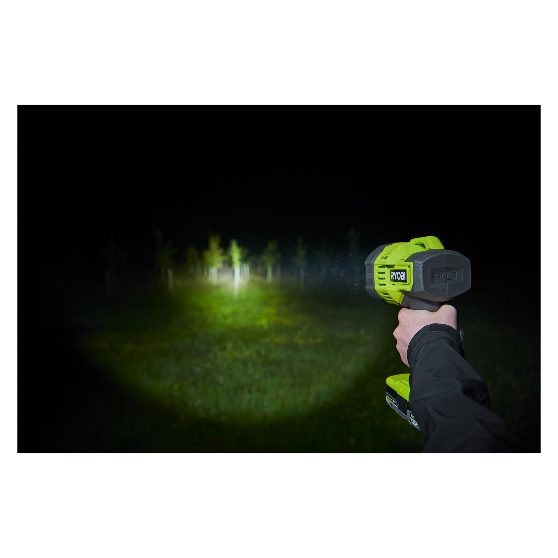 En person bär en Ryobi One+ portabel LED-strålkastare RLS18-0, som lyser med en stark ljusskena som lyser upp ett runt område med gräs och träd i mörkret utomhus på natten.