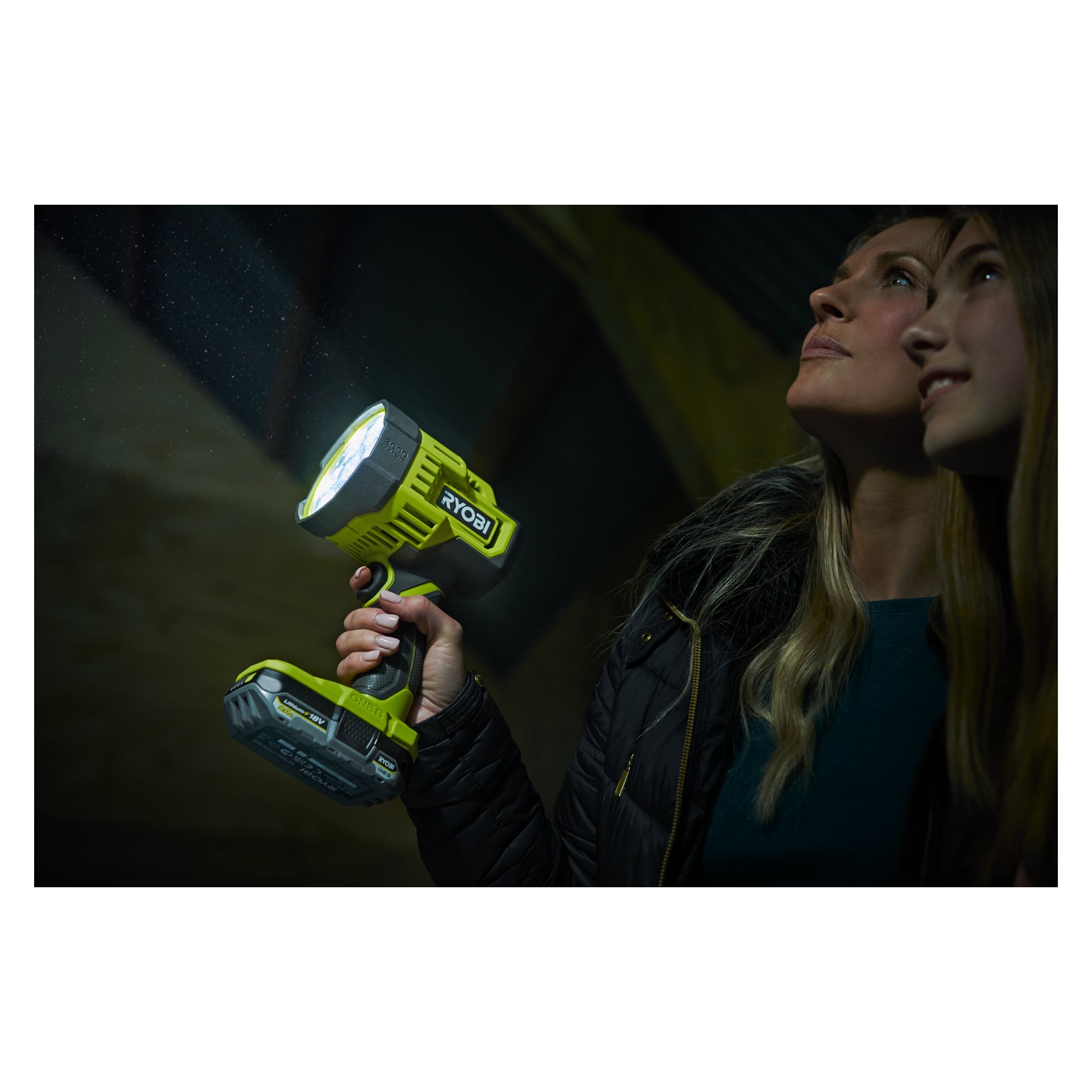 I ett svagt upplyst utrymme tittar två personer i jackor uppåt när en av dem håller en skinande Ryobi One+ Portable LED-pannlampa RLS18-0. Strålen lyfter fram flytande dammpartiklar, och båda individerna verkar undersöka något ovanför.
