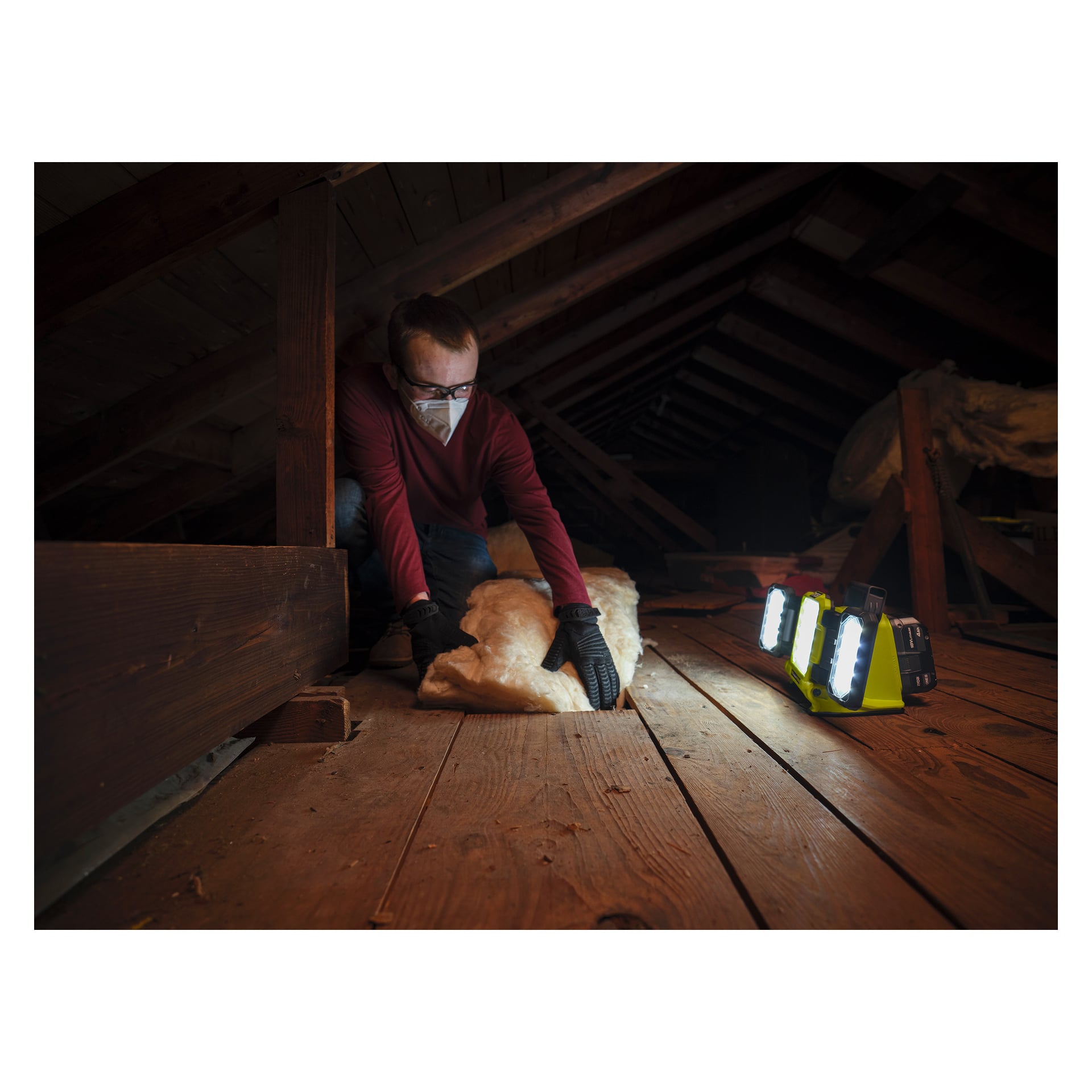 En person klädd i mask och handskar installerar isolering på en vind med svagt ljus, upplyst av Ryobi One+ batteridrivna LED-arbetslampa RLP18-0, som kastar ljus på trägolvet och balkarna.