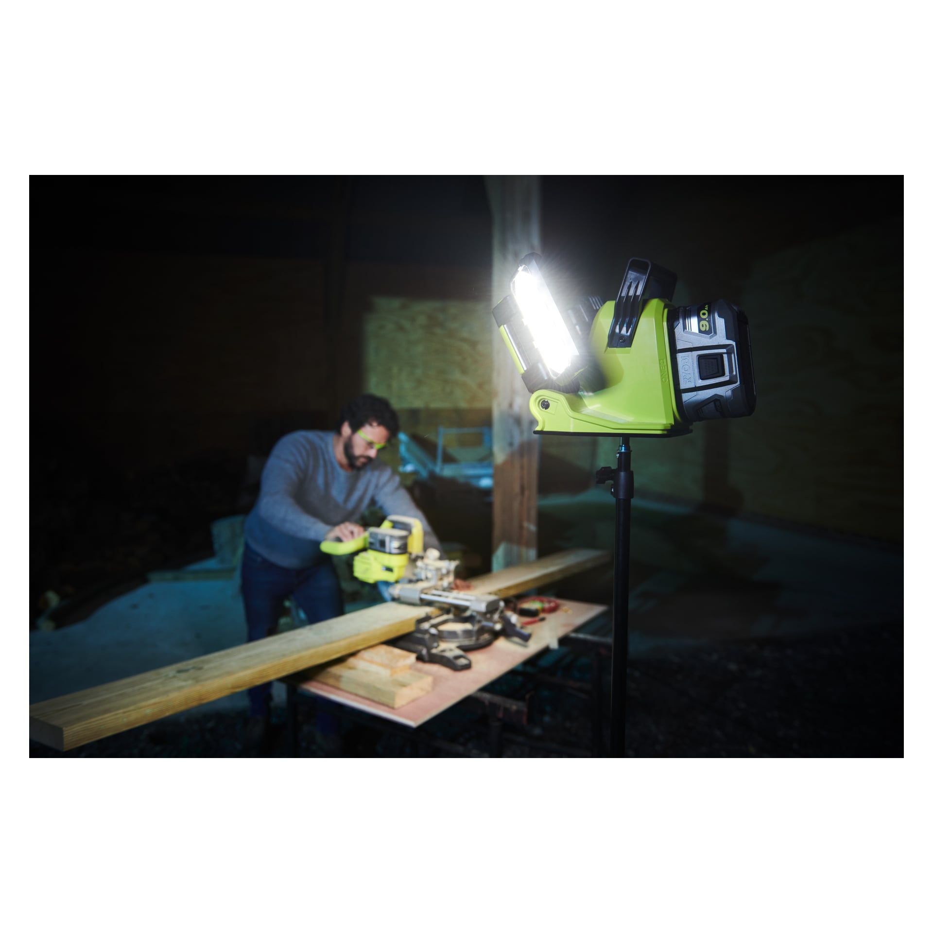 I en svagt upplyst verkstad använder någon en såg för att såga en träplanka på en arbetsbänk. En starkt lysande Ryobi One+ batteridriven LED-arbetslampa (RLP18-0) på ett stativ lyser upp området framför.
