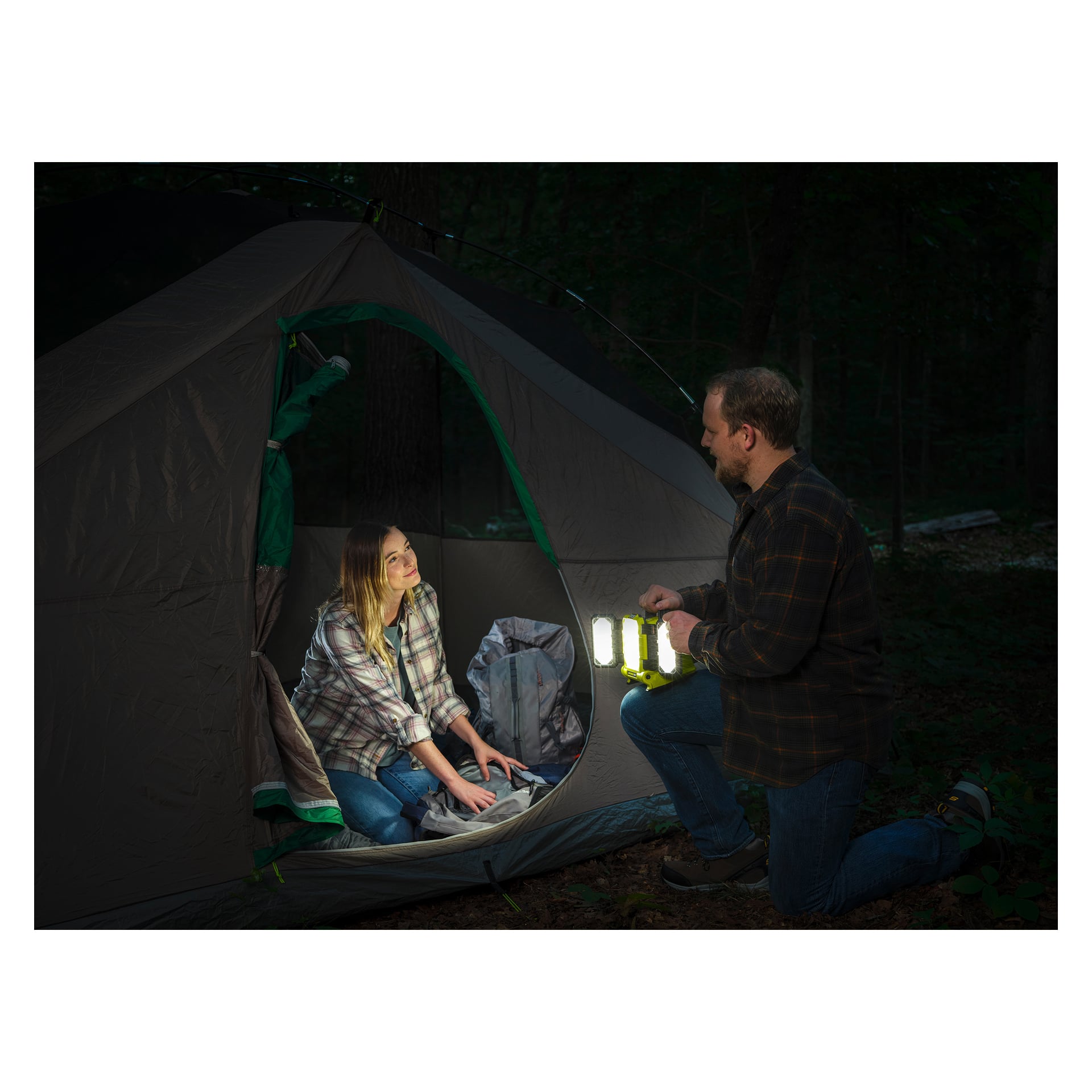 En kvinna sitter inne i ett tält och tittar på en man som knäböjer utanför och håller i en upplyst Ryobi One+ batteridriven LED-arbetslampa (RLP18-0). Båda är klädda i rutiga skjortor. Scenen utspelar sig på natten i ett skogsområde, med olika campingutrustning synliga inne i tältet.