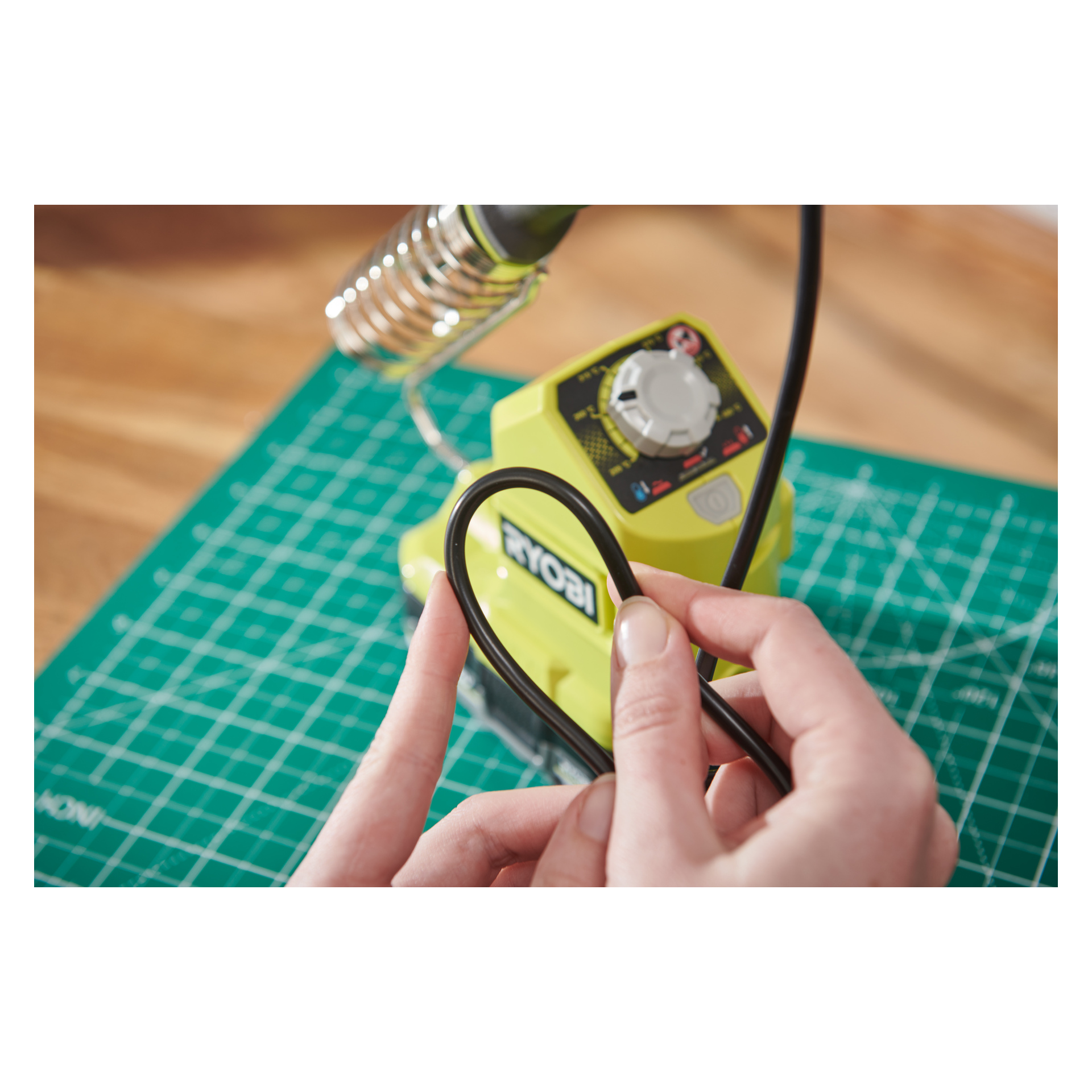 En person håller en svart kabel nära en Ryobi One+ batteridriven lödkolv RSI18-0, placerad på en grön skärmatta märkt med mätrutor. Lödkolven sitter säkert i sin hållare och enhetens kontrollratt syns.