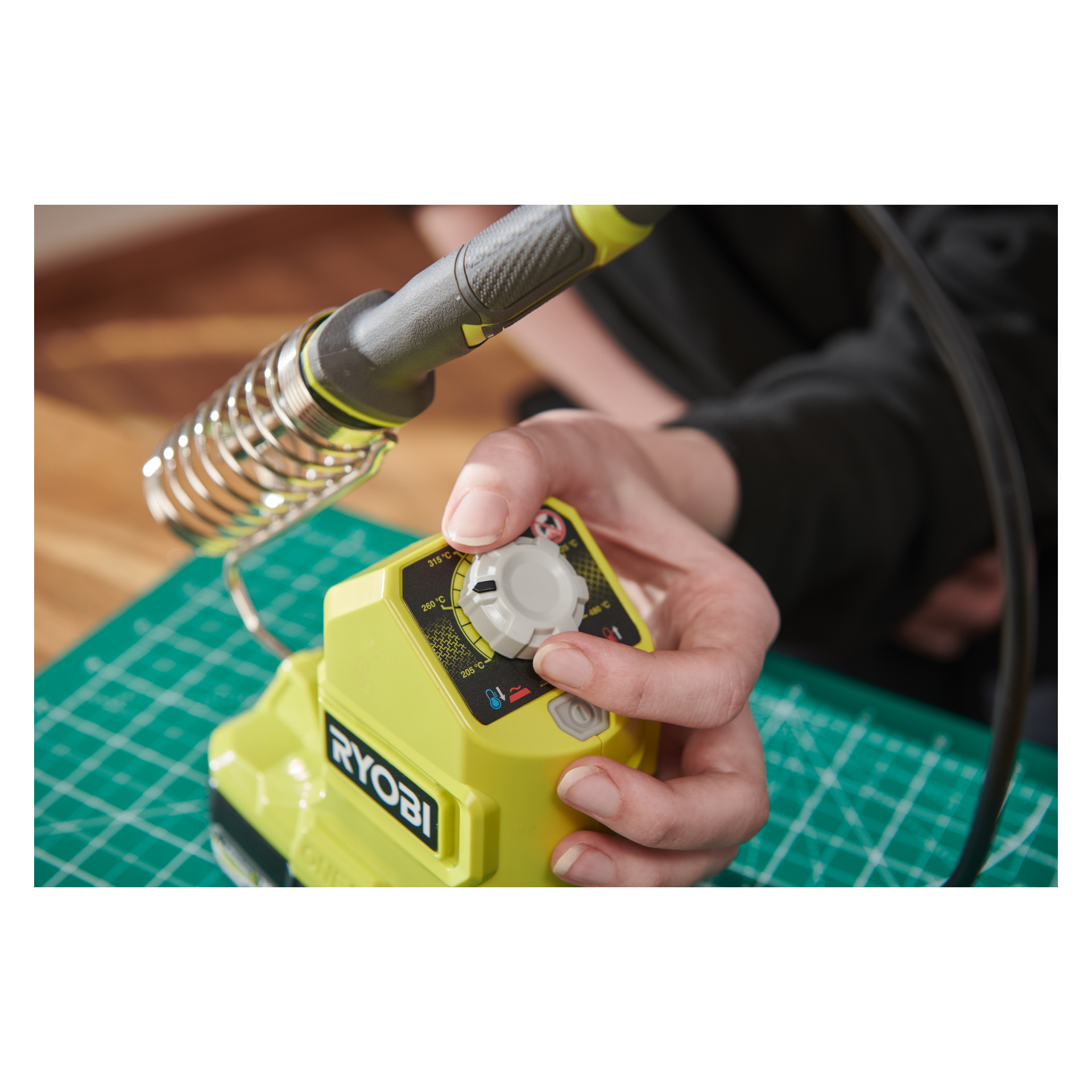 En person justerar temperaturratten på en Ryobi One+ batteridriven lödkolv RSI18-0 med höger hand på en grön skärmatta. Lödkolven sitter säkert i sin hållare när de vrider på ratten.