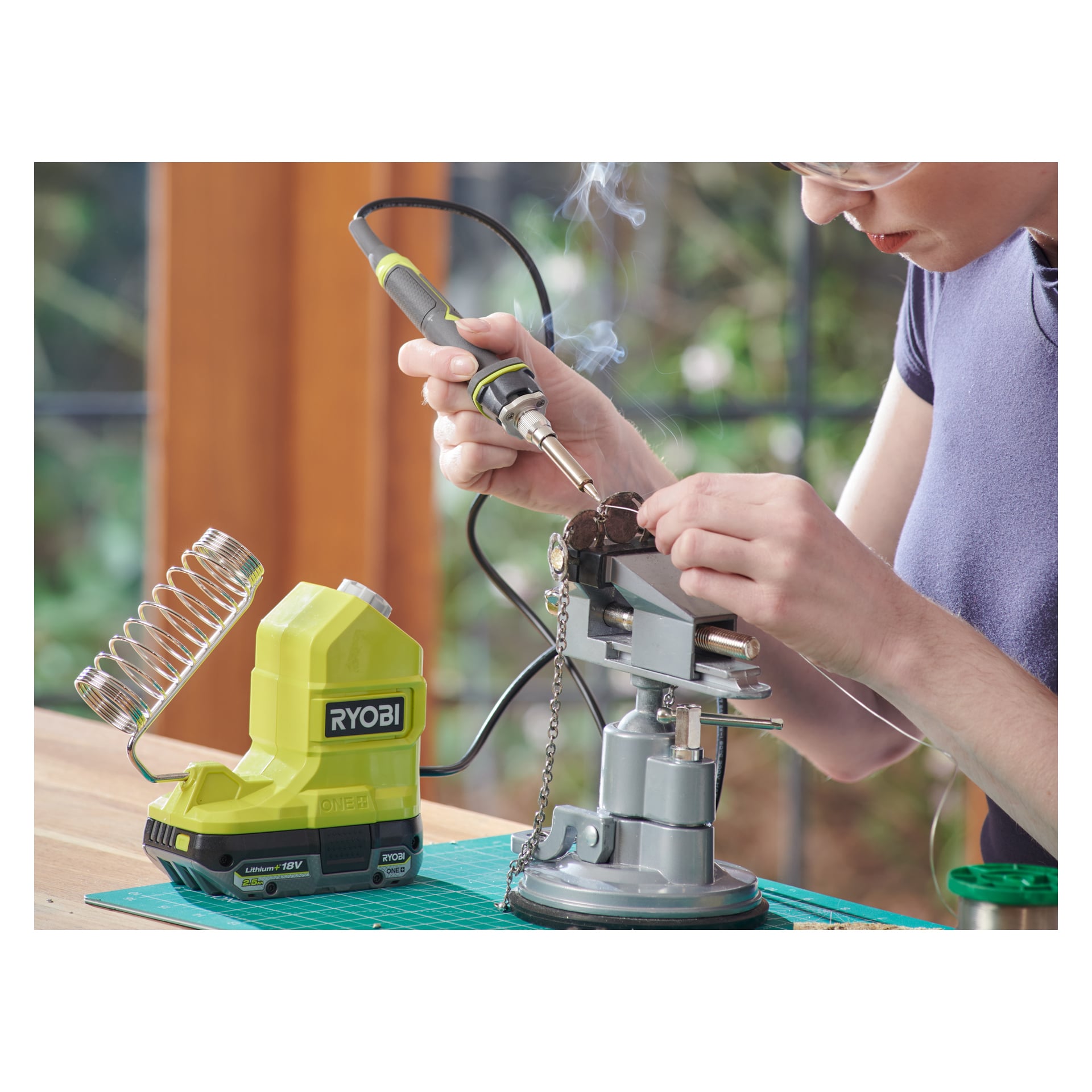 Någon använder en batteridriven Ryobi One+ RSI18-0 lödkolv för att arbeta med en liten elektronisk del som hålls i ett skruvstäd. Den gröna Ryobi One+ lödstationen placeras i närheten på bordet, bredvid en skärmatta, och rök stiger från lödkolvens spets.
