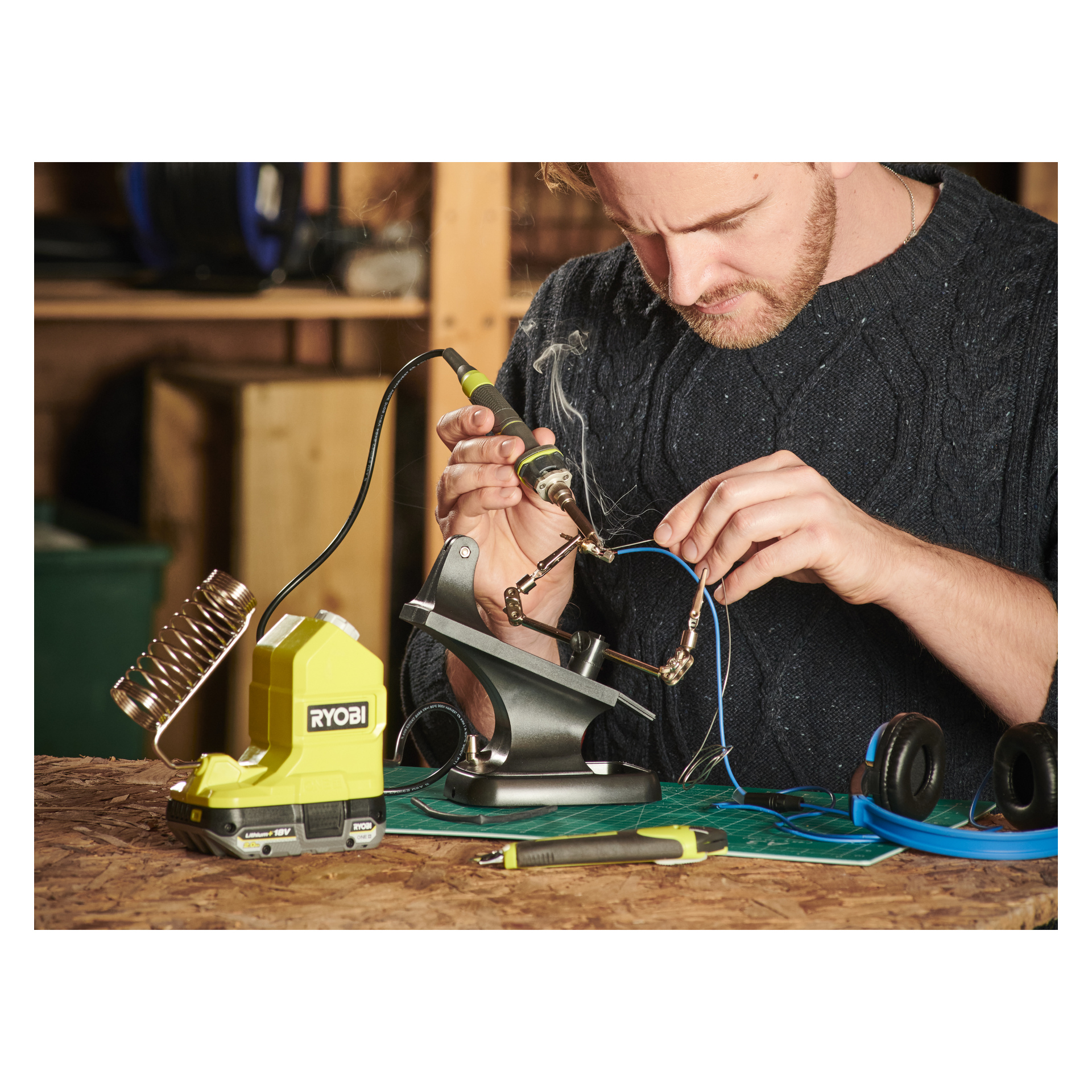 Någon monterar elektronik vid en arbetsbänk med en Ryobi One+ batteridriven lödkolv RSI18-0. Blå kablar och blå hörlurar är också ett alternativ.