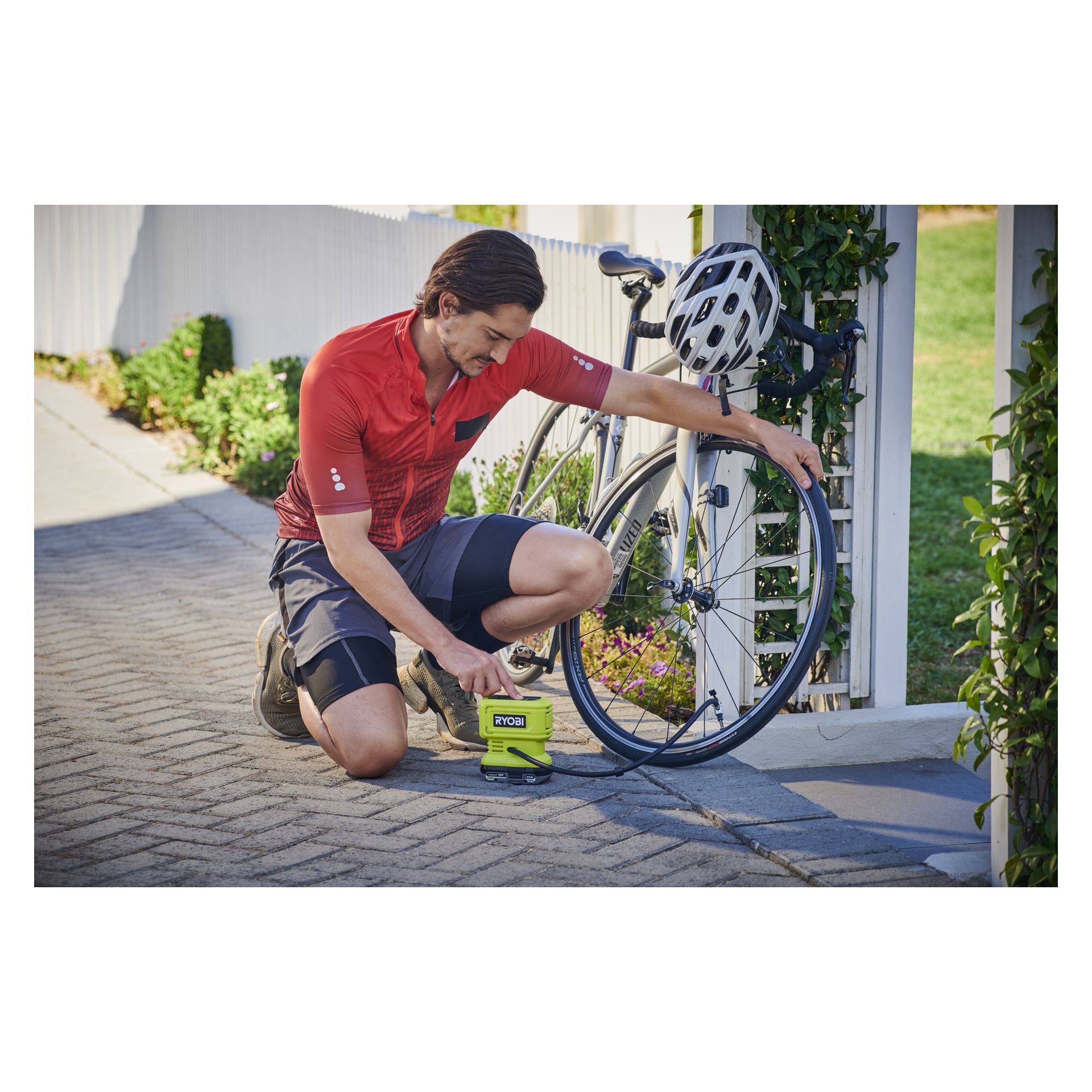 Mężczyzna w czerwonej koszulce rowerowej klęka na chodniku, pompując przednią oponę roweru szosowego za pomocą mini pompy powietrza Ryobi One+ RPI18-0 zasilanej baterią. Na kierownicy motocykla umieszczony jest biały hełm.