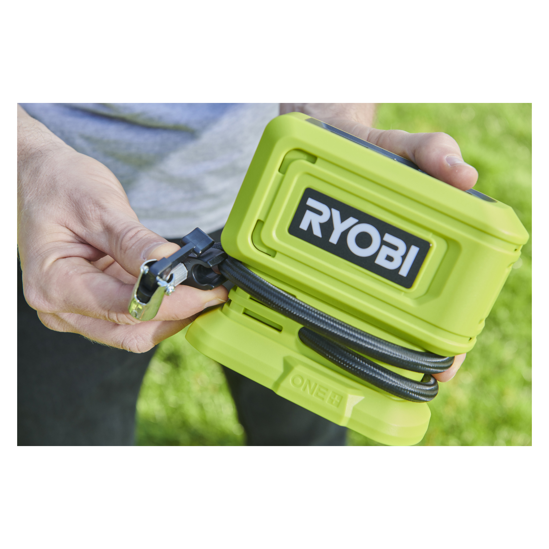 Ktoś na zewnątrz trzyma zieloną mini pompę powietrza Ryobi One+ zasilaną baterią (model RPI18-0), z widocznym podłączonym wężem i dyszą powietrza. Na przodzie pompy widnieje logo Ryobi, a w tle widać trawę.