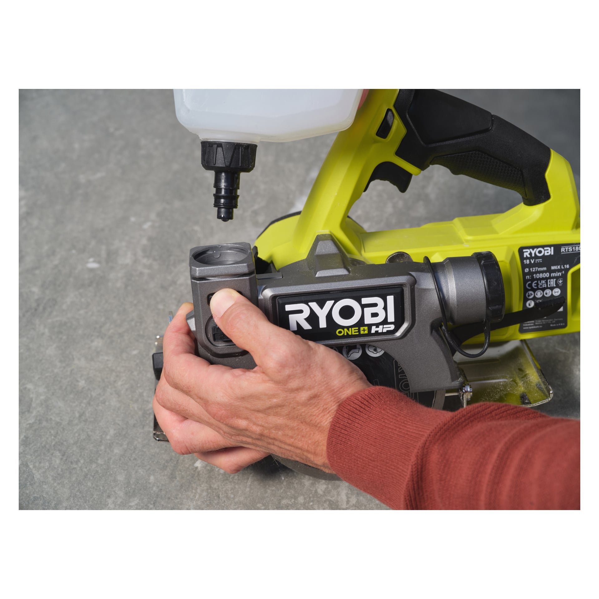 En hand justerar en komponent på en Ryobi One+ HP batteridriven platta och klinksåg RTS18C-0 som vilar på en grå yta. Sågen har en gul och grå kropp med en exponerad vattentank.