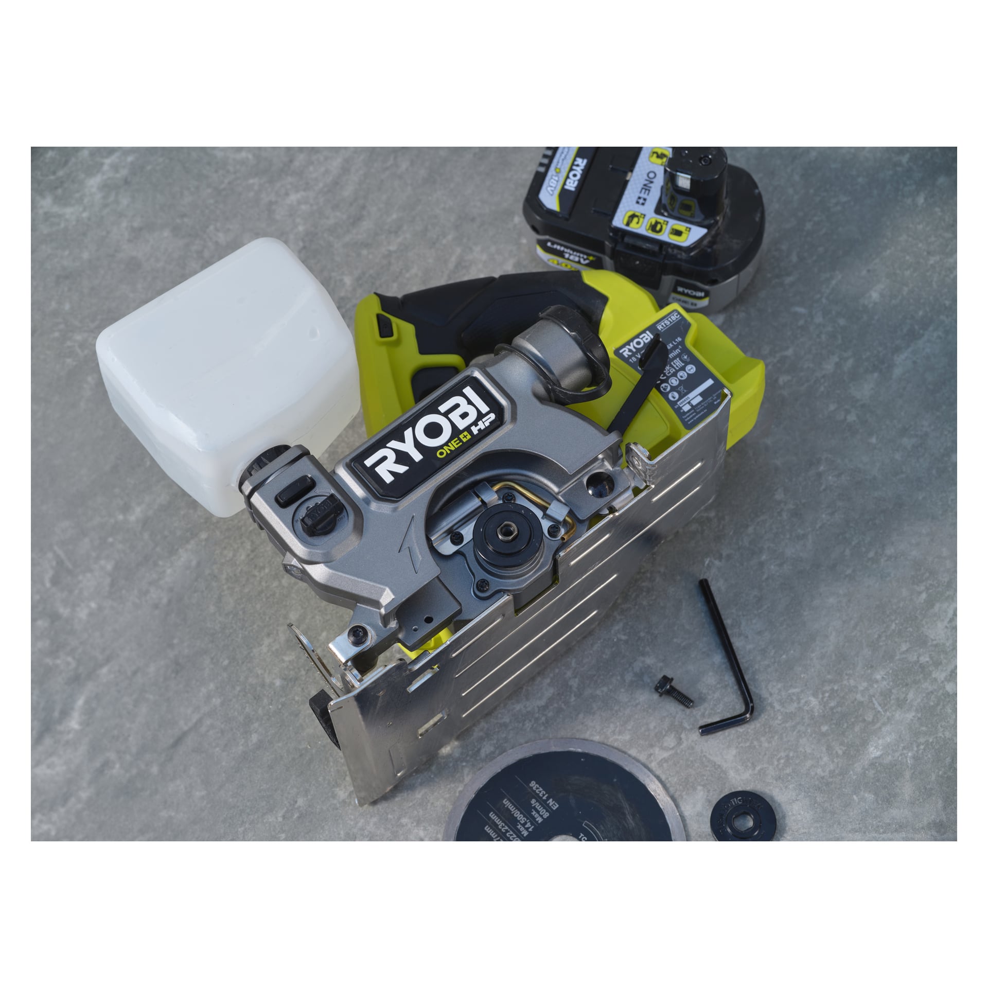 En Ryobi One+ HP batteridriven platte- och klinksåg RTS18C-0 visas på en grå yta, tillsammans med en vit behållare, ett batteripaket, en metallskiva och en insexnyckel. Sågen är delvis isär, vilket blottar några av dess inre delar.