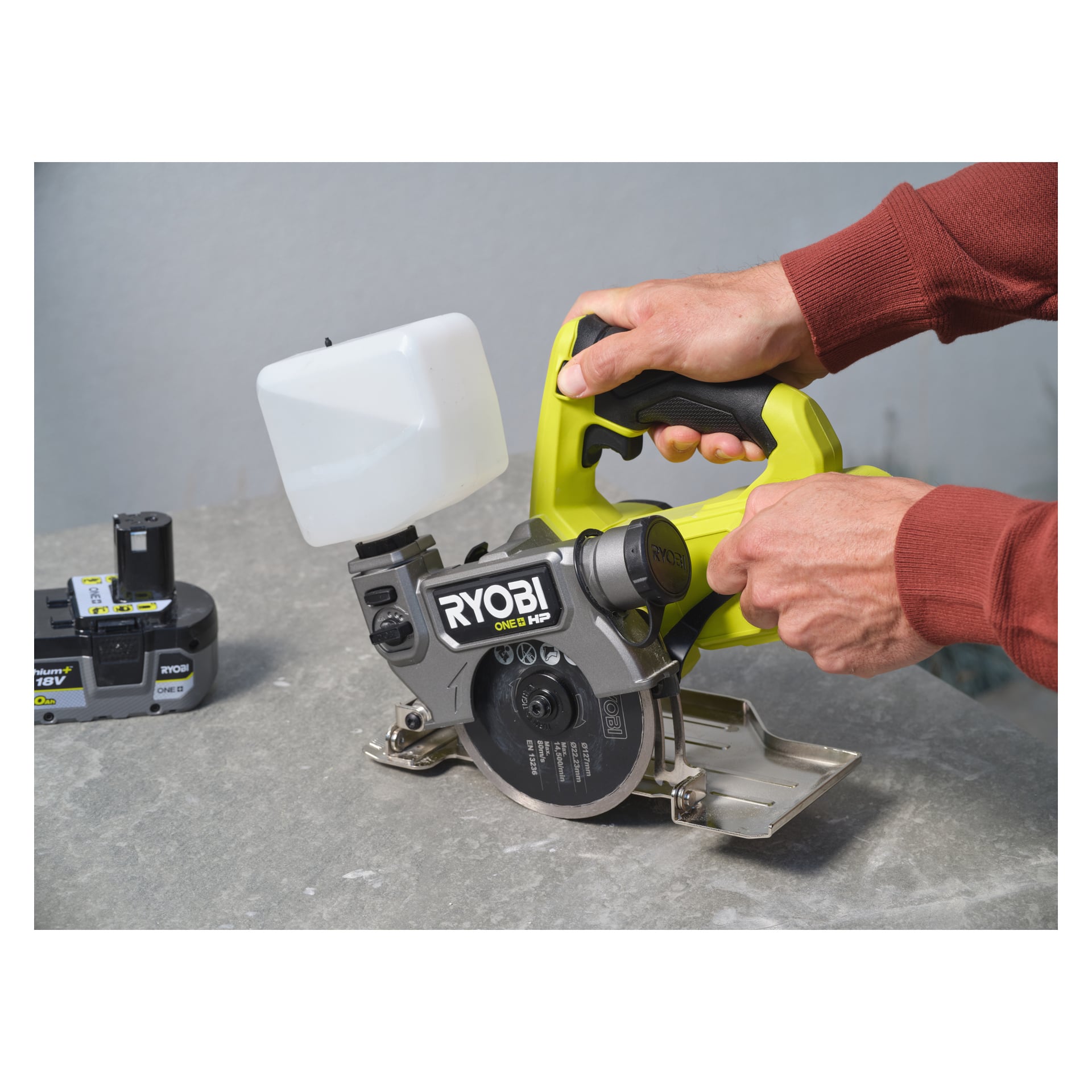 Någon som bär en röd tröja håller i en Ryobi One+ HP batteridriven platta och klinksåg RTS18C-0, som har en vattentank och vilar på en grå betongyta. Ett 18V Ryobi-batteri ligger i närheten.
