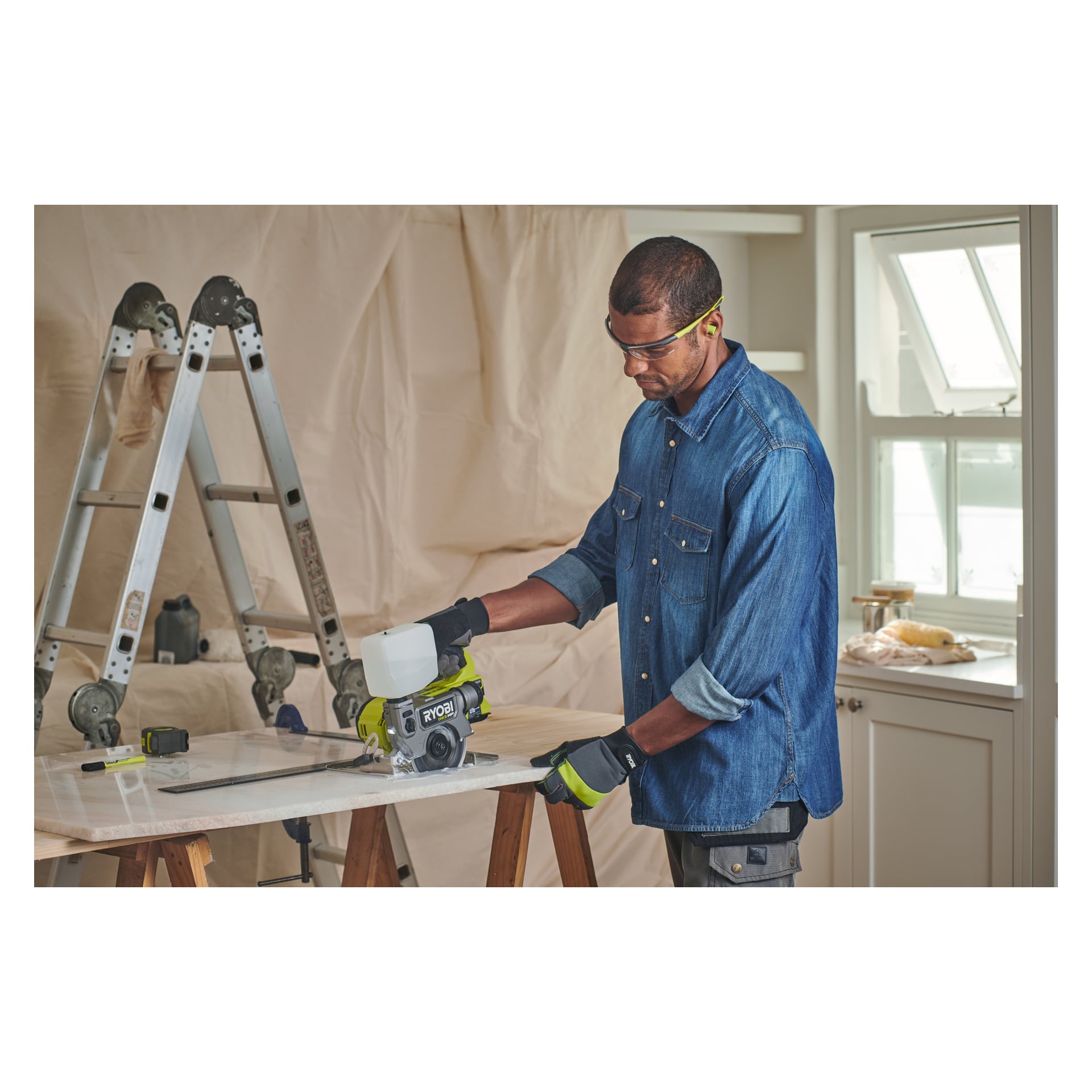 En man i skyddsglasögon och handskar använder en Ryobi One+ HP batteridriven kakel och klinksåg (RTS18C-0) för att såga en träbit inomhus. Bakom honom finns en stege, hyllor täckta med skyddsmaterial och ett fönster.