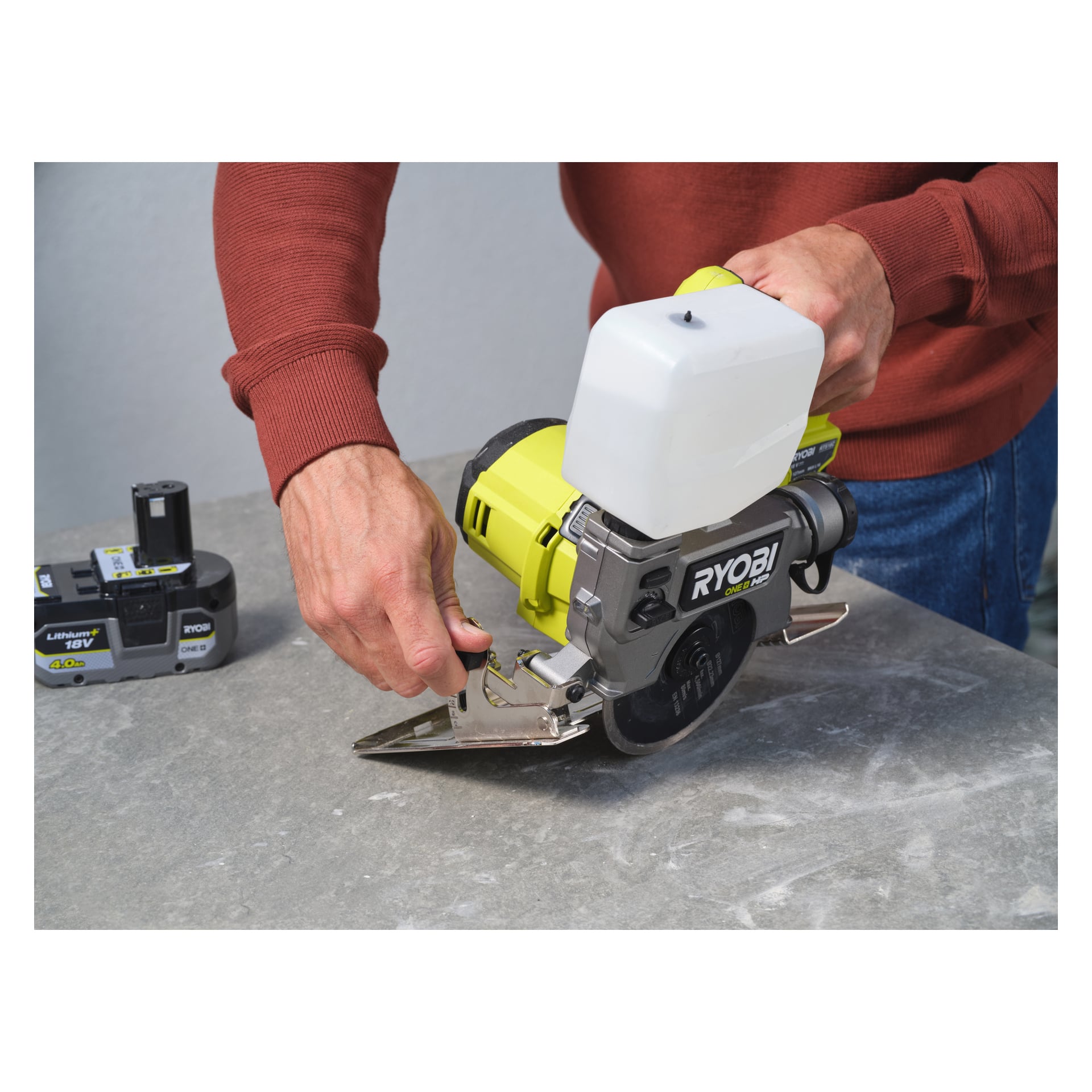 Iklädd en röd tröja justerar man en Ryobi One+ HP batteridriven platta och klinksåg RTS18C-0 placerad på en grå yta. Till vänster sitter ett separat Ryobi 18V-batteri. Personens händer är nära bladskyddet på sågen.