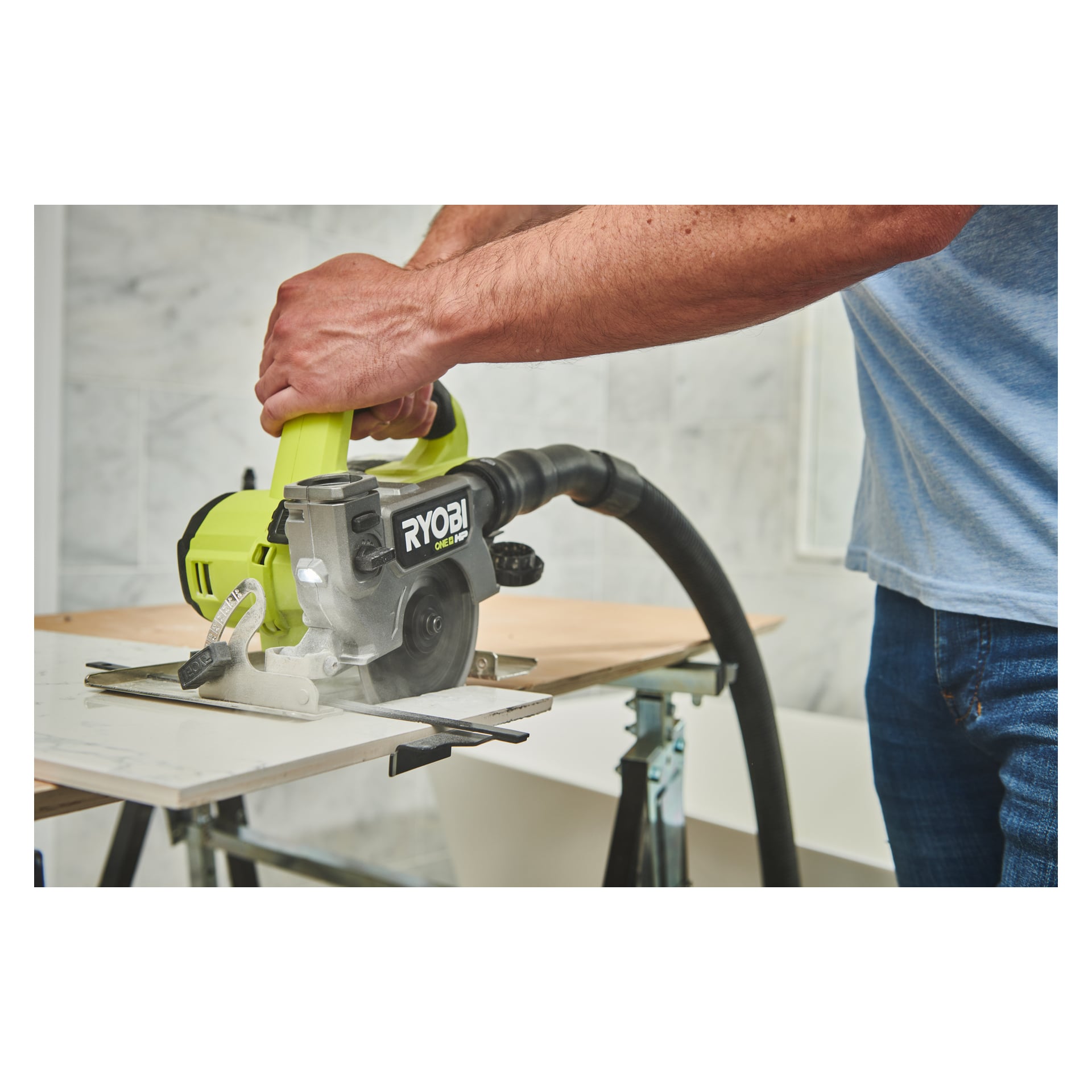 Någon använder en Ryobi One+ HP batteridriven kakel och klinksåg (RTS18C-0) för att såga igenom en platta, med en vakuummontering kopplad till verktyget. Arbetsplatsen har en marmormönstrad vägg i bakgrunden.