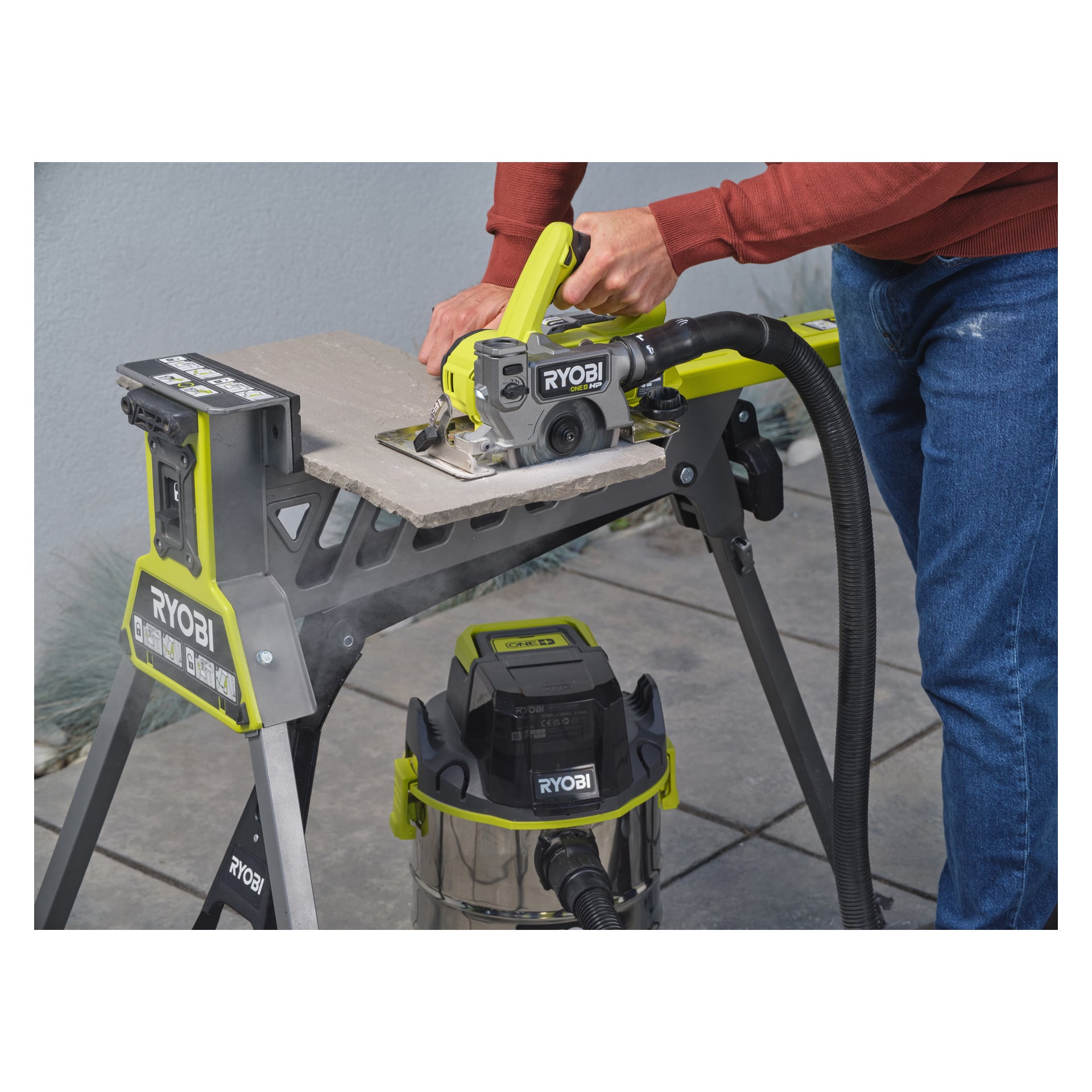 En person använder en Ryobi One+ HP batteridriven platta och klinksåg RTS18C-0 utrustad med vakuumtillbehör för att såga en kakel på en Ryobi-arbetsbänk. Scenen utspelar sig utomhus på kaklade golv, med grönska i bakgrunden.