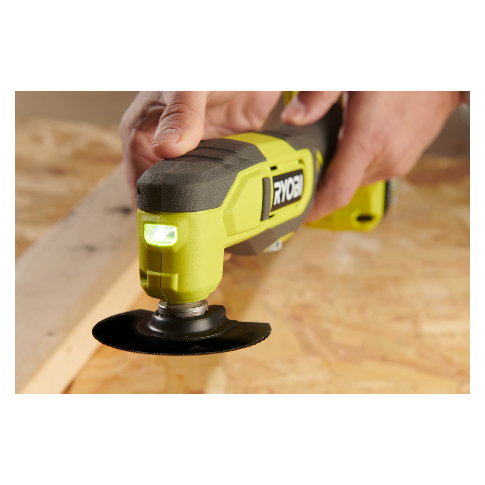 Någon håller i en grön Ryobi One+ Multitool RMT18-0, utrustad med ett cirkulärt blad, nära en träbit som vilar på en arbetsbänk.