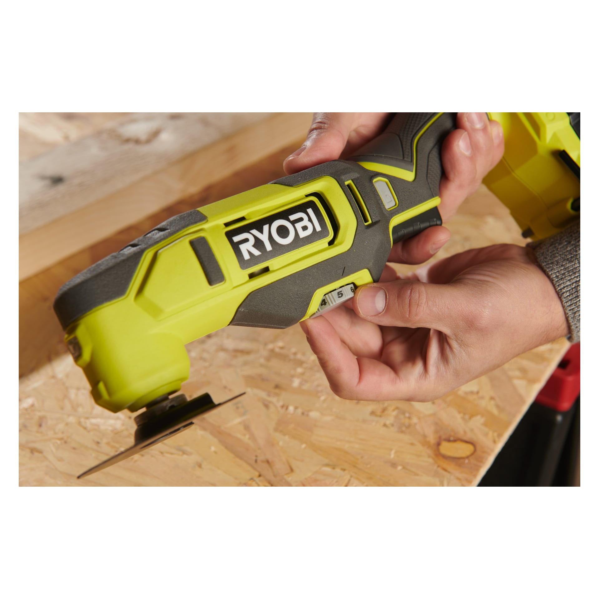 Någon justerar kontrollerna på en grön Ryobi One+ Multitool RMT18-0, håller den ovanför en träyta där en del av verktyget vilar mjukt mot träet.