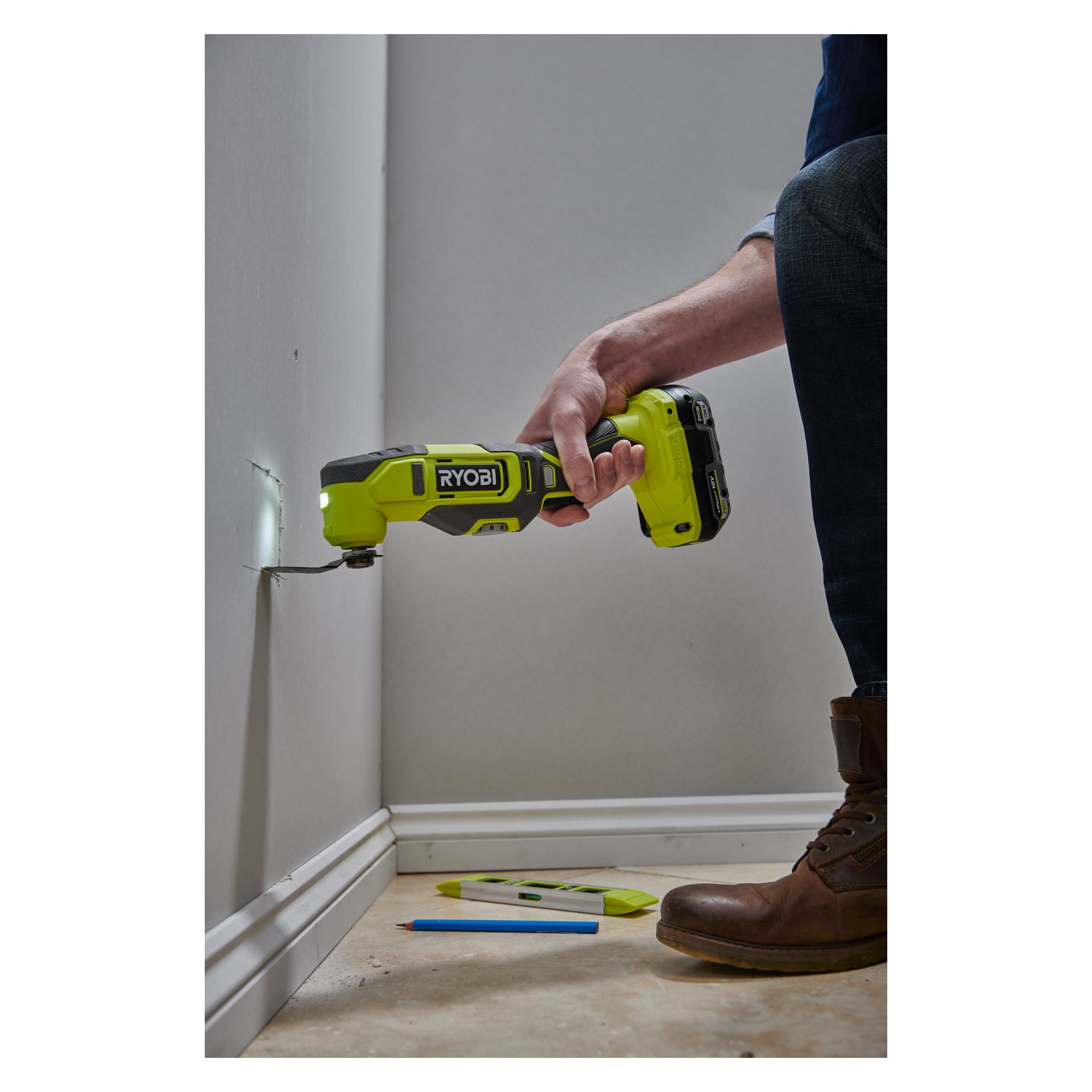 En person använder en grön Ryobi One+ Multi-tool RMT18-0 för att skära ett hål i en vit vägg nära golvet, med en penna och mätverktyg liggande i närheten.