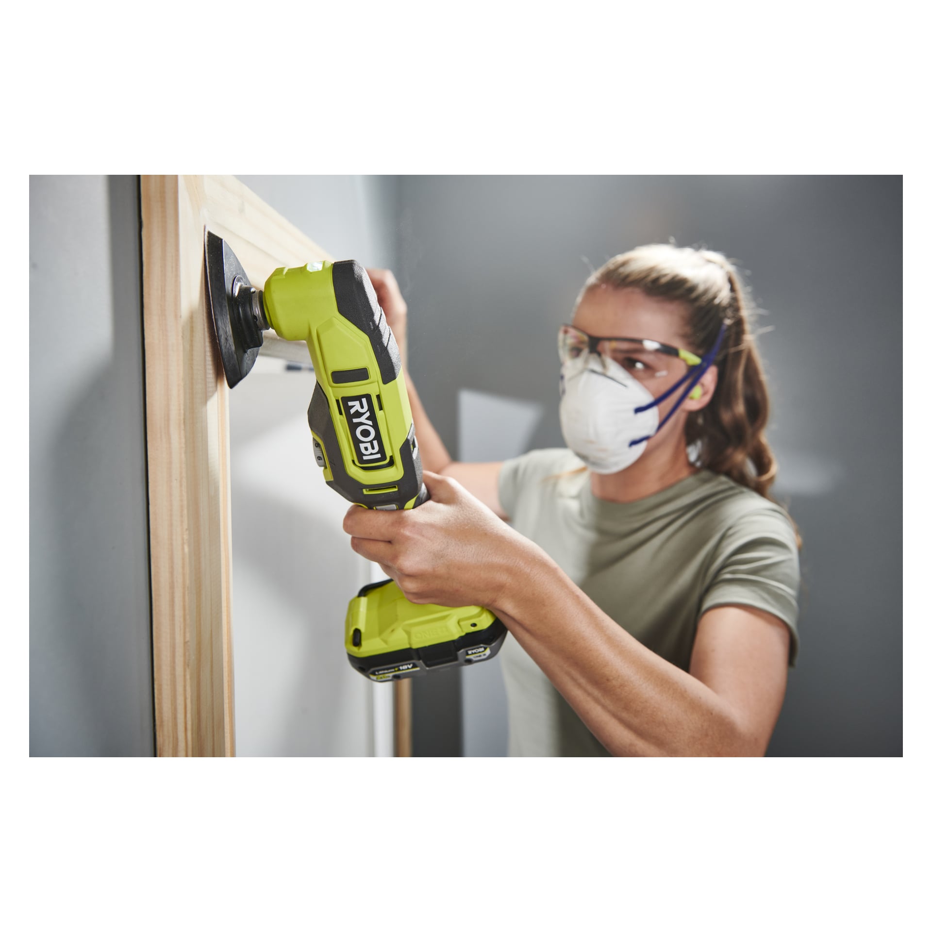 En person utrustad med skyddsglasögon och mask använder Ryobi One+ Multi-tool RMT18-0 för att slipa eller jämna ut kanten på en träram som står mot en vägg.
