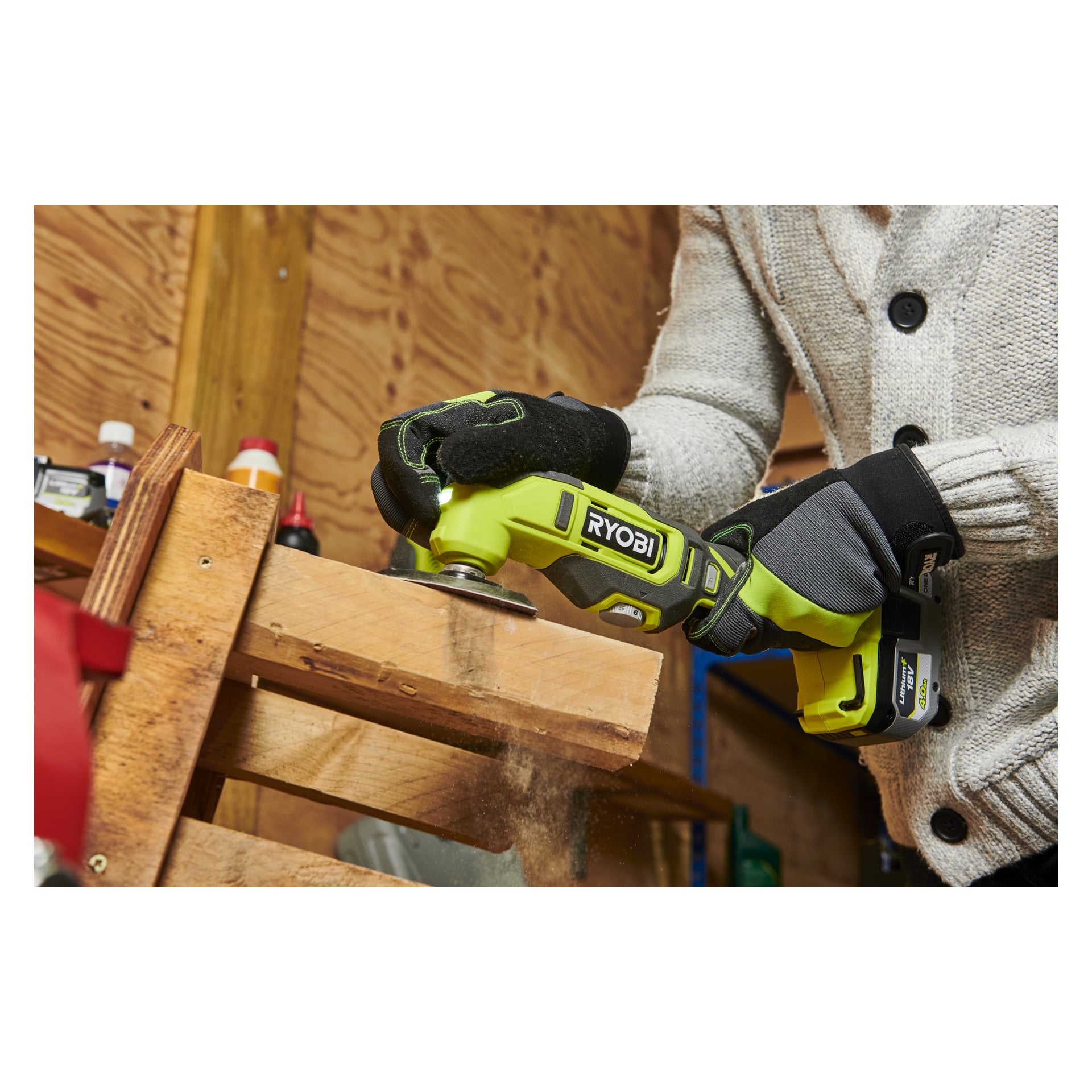 En person med handske slipar en träplanka i en verkstad med hjälp av en Ryobi One+ Multitool RMT18-0. Sågspån svävar i luften, och hyllor fyllda med olika föremål kan ses bakom dem.