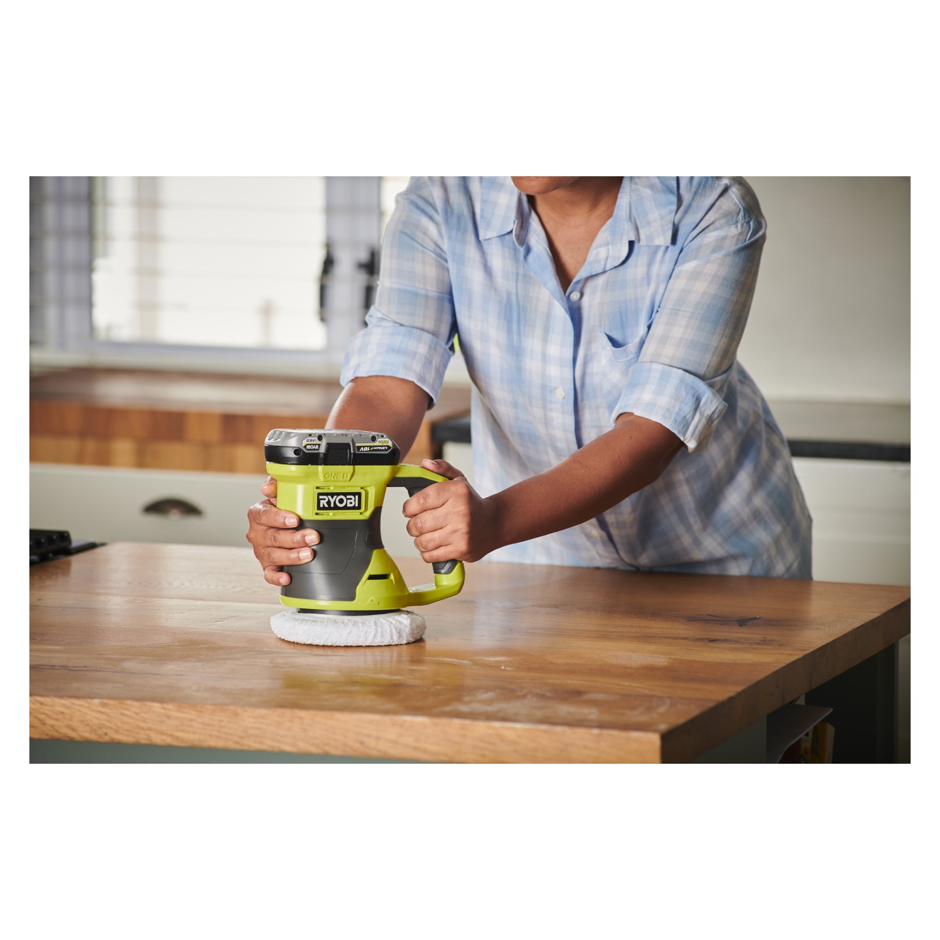 Iklädd en rutig skjorta använder någon en grön och svart Ryobi One+ Oscillating Polishing Machine RBP18150-0 för att polera en träbänkskiva i ett ljust kök med fönster och synliga köksytor.
