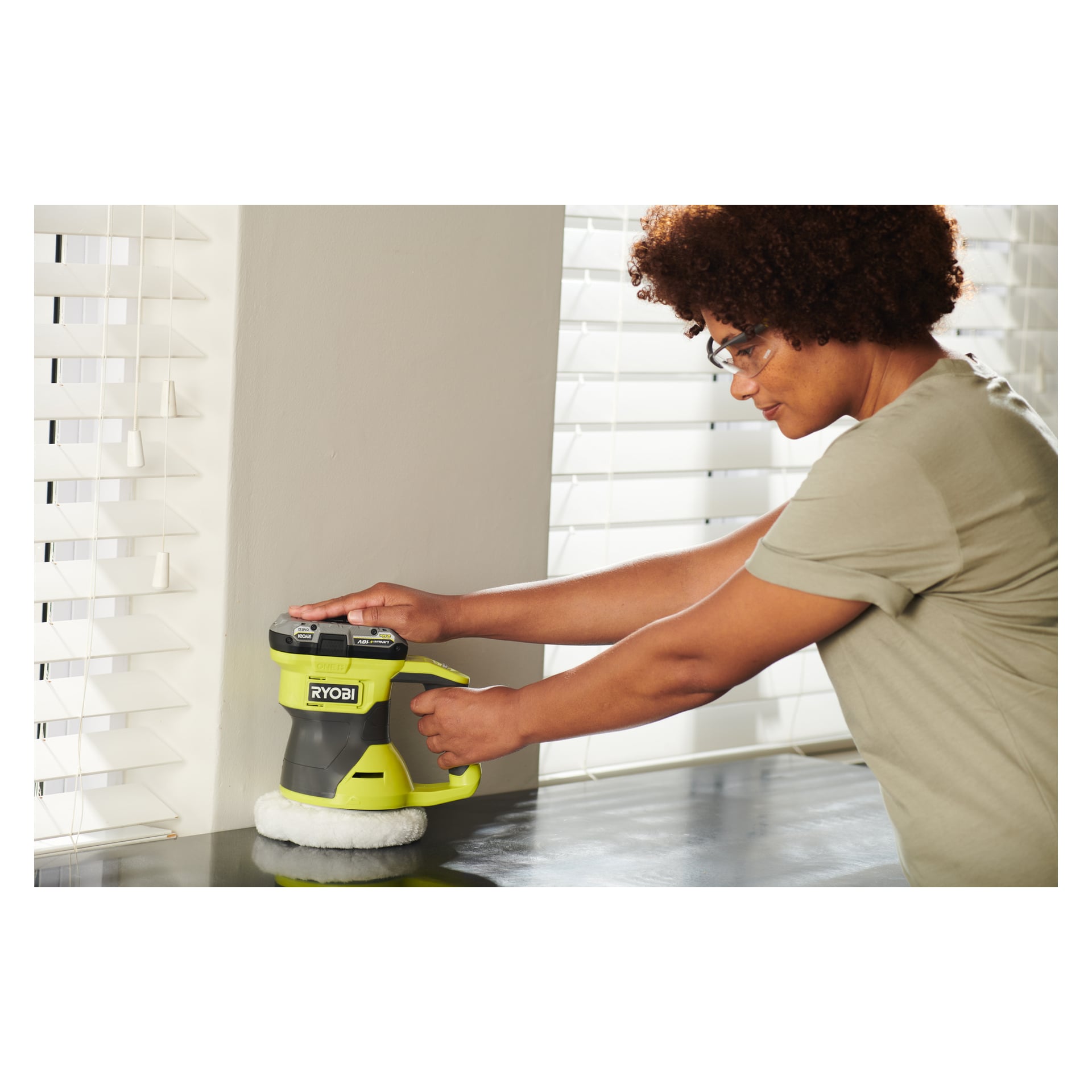 En person som bär skyddsglasögon och en ljus skjorta använder en Ryobi One+ Oscillating Poleringsmaskin RBP18150-0 på en mörk bänkskiva bredvid vita persienner.