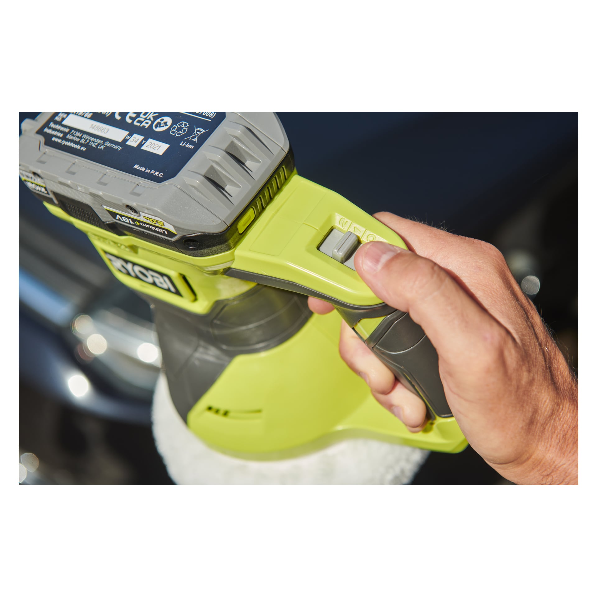 En närbild visar en hand som greppar handtaget och kontrollbrytaren på Ryobi One+ Oscillating Polishing Machine RBP18150-0, med en suddig bil delvis synlig i bakgrunden.