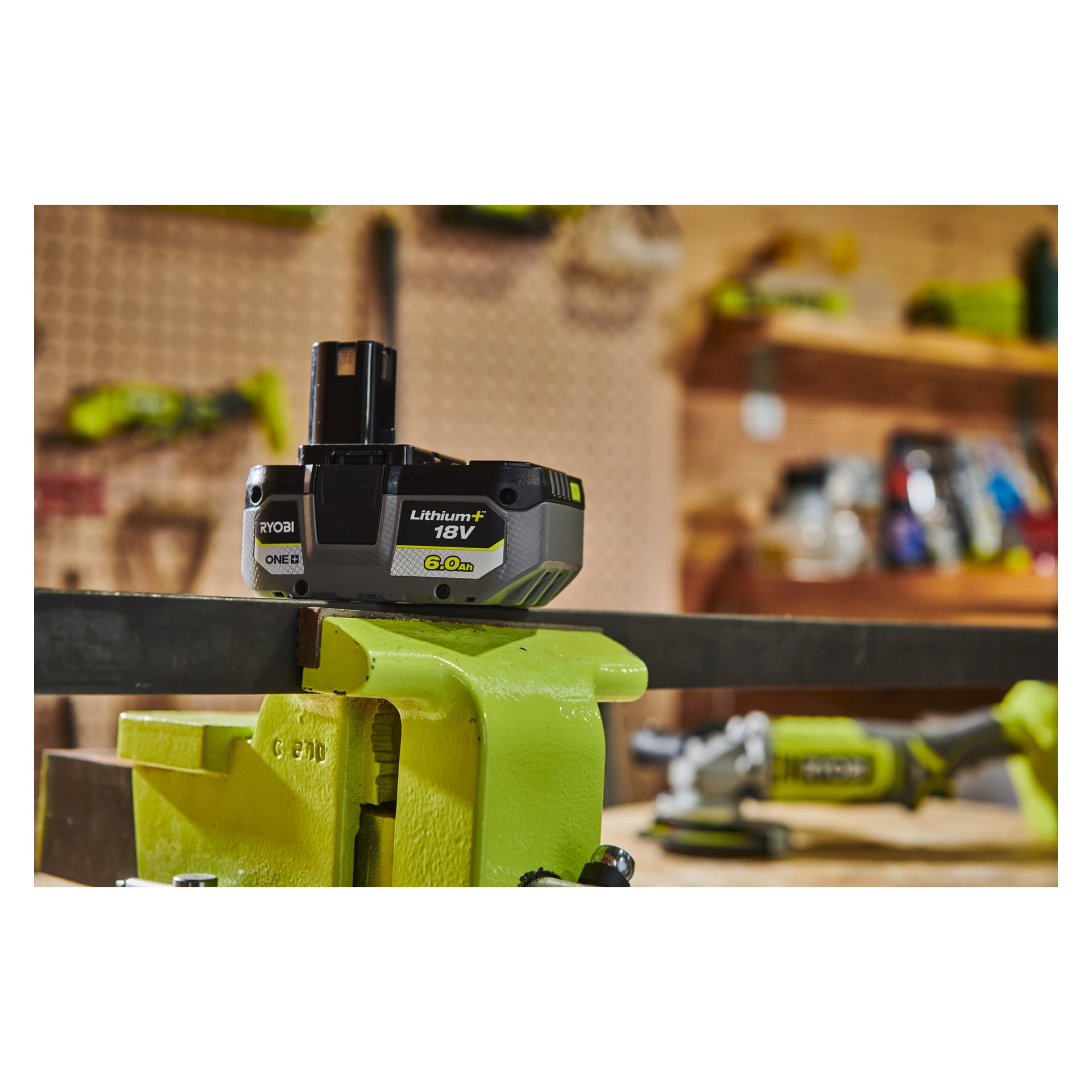 "Ryobi One+ Battery RB1860X 18V 6.0Ah" stoi na metalowym prętze trzymanym przez zielone imadło w warsztacie, z rozmazanymi narzędziami i półkami za nimi.