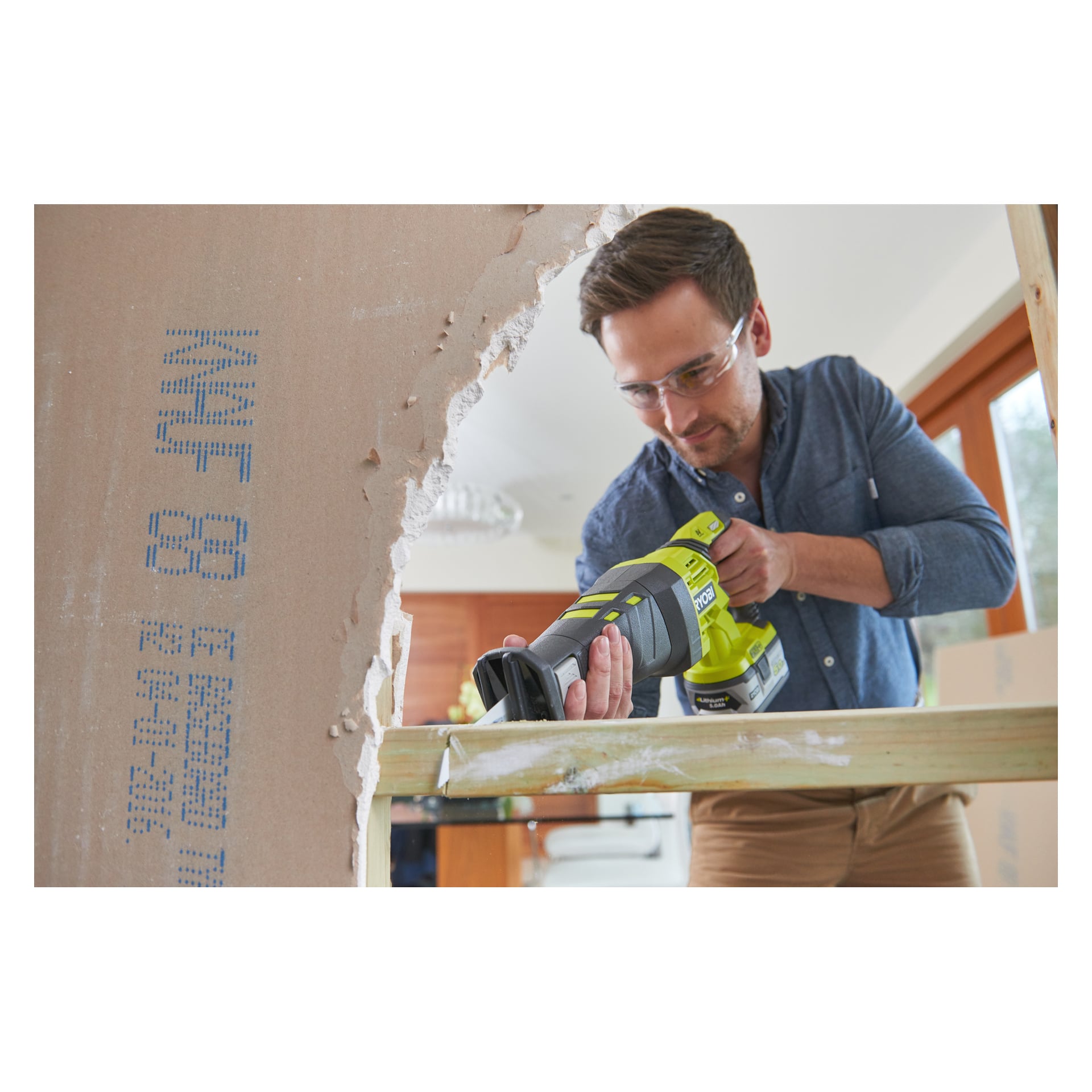 Mit Schutzbrille bedient man eine grün-schwarze Ryobi One+ batteriebetriebene Kolbensäge (R18RS-0), um Holz innerhalb des Rahmens einer teilweise abgerissenen Innenwand zu schneiden.
