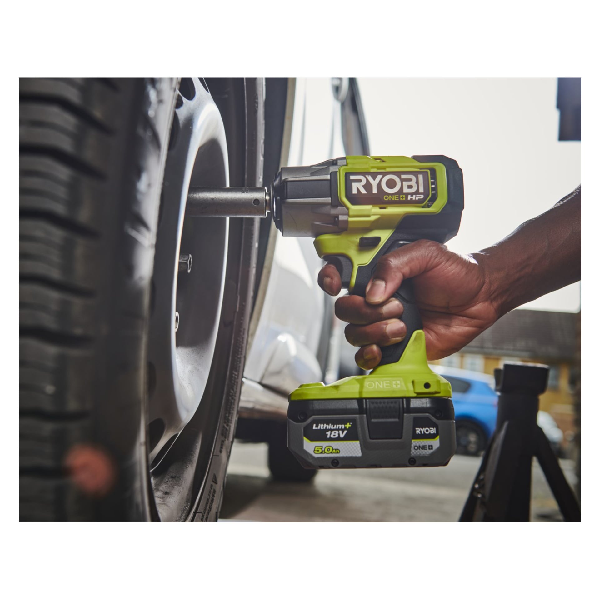 Jemand bedient den Ryobi One+ HP Schlagschrauber und Mini-Kompressor (RIW18XPI-0 | R18PI-0, Satz aus zwei Modellen), um eine Radmutter an einem Autorad zu verstellen, während ein Wagenheber das Auto dahinter hält.