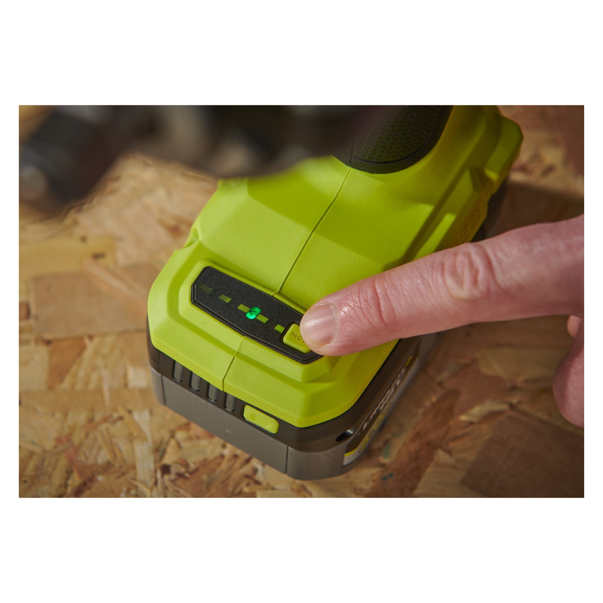 Käsi painaa nappia Ryobi One+ HP -iskuavaimessa ja minikompressoriparistossa, jolloin neljä osittain valaistua vihreää LED-vilkkua lepäävät puupinnalla.