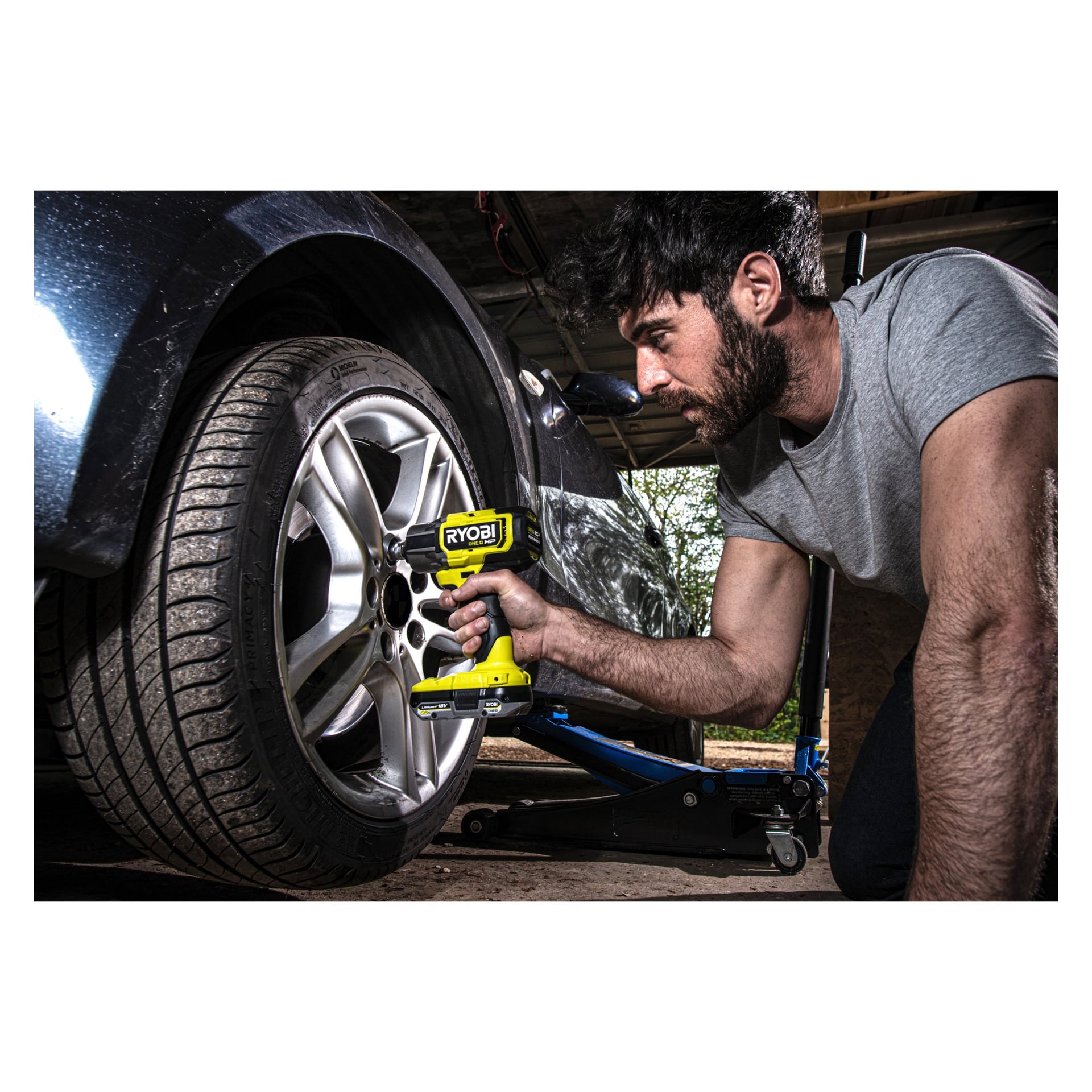 Mies käyttää Ryobi One+ HP -iskuavainta ja Mini Compressoria RIW18XPI-0 | R18PI-0 (2 kappaleen sarja) auton pyörän muttereiden käsittelyyn, ja ajoneuvo nostetaan sinisellä hydraulisella lattiatunkilla autotallissa tai työpajassa.