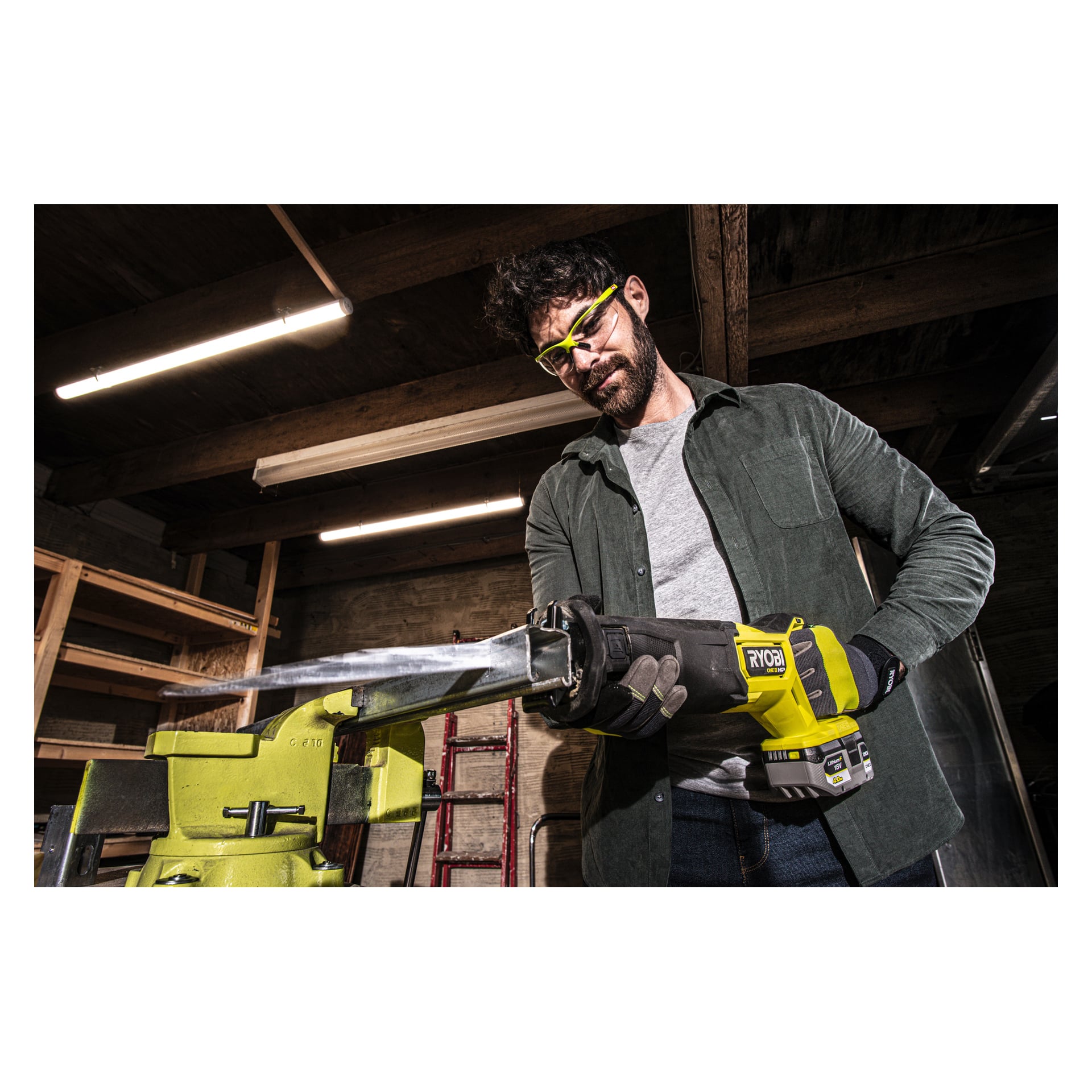 Eine Person, die Schutzbrille und Handschuhe trägt, bedient eine gelb-schwarze Ryobi One+ HP bürstenlose Hubkolbensäge RRS18X-0, die ein Metallrohr durchschneidet, das von einem grünen Schraubstock in einer Werkstatt mit Holzregalen gehalten und von Leuchtstoffröhren beleuchtet wird.