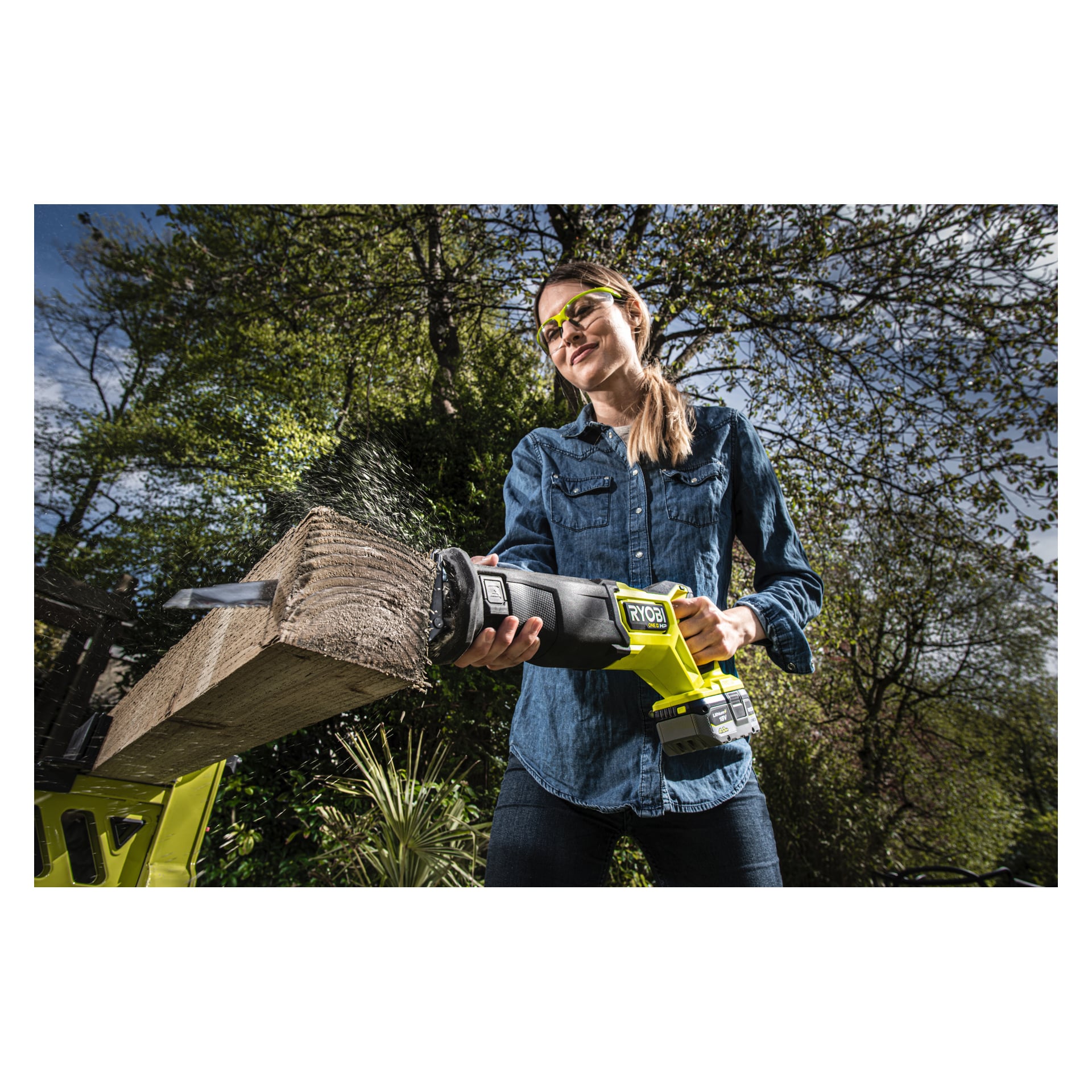 Mit Schutzbrille benutzt man eine Ryobi One+ HP batteriebetriebene bürstenlose Kolbensäge RRS18X-0, um draußen durch ein dickes Holzbrett zu schneiden, wobei Holzspäne in die Luft sprühen, während Bäume und Pflanzen dahinter sichtbar sind.