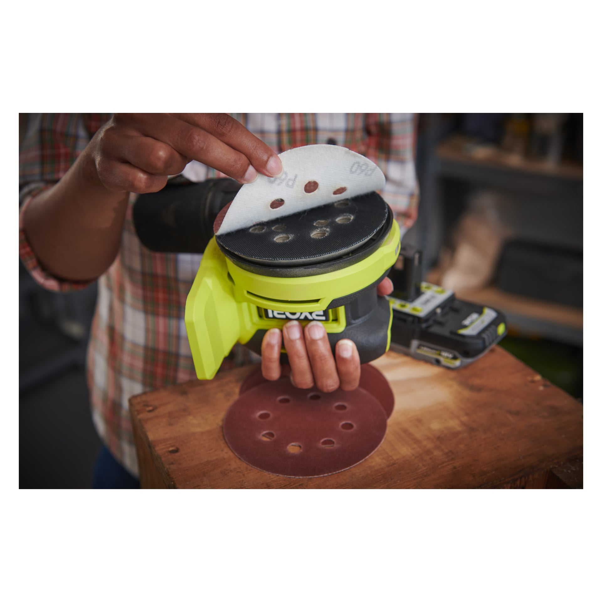 In einer Werkstatt montiert eine Person im karierten Hemd eine runde Schleifscheibe auf den Ryobi One+ HP Eccentric-Schleifer RROS18C-0, während mehrere andere Schleifscheiben auf der darunterliegenden Holzoberfläche verteilt sind.